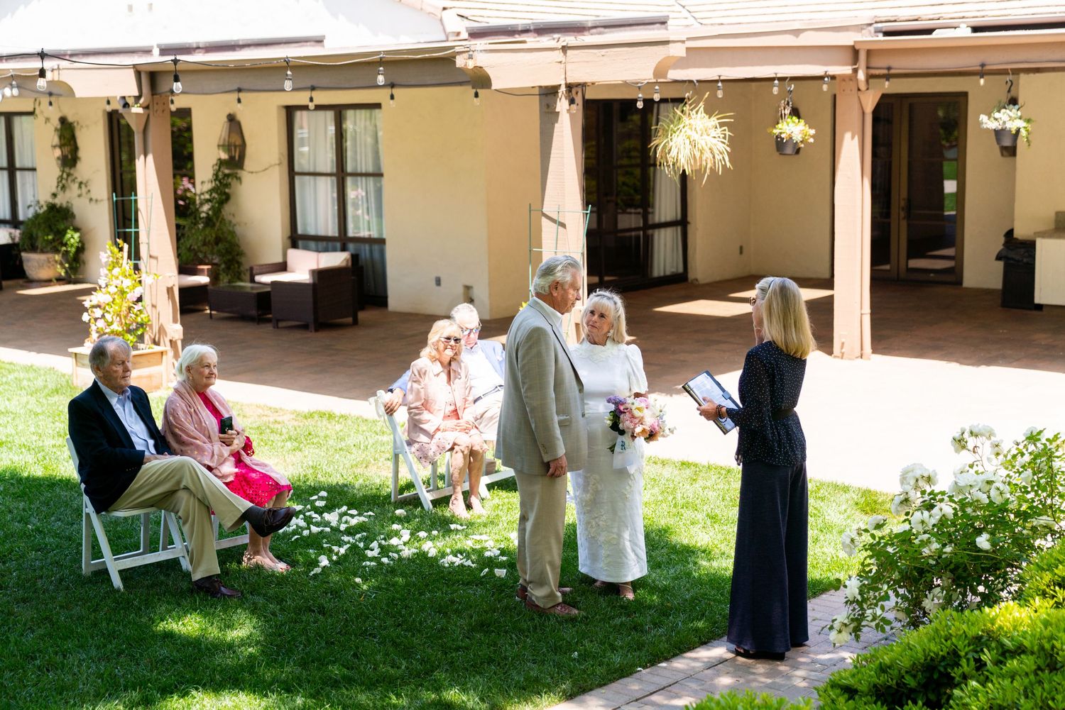 Guests socialize on a green lawn outside a Spanish-style building with covered patio and hanging lights.