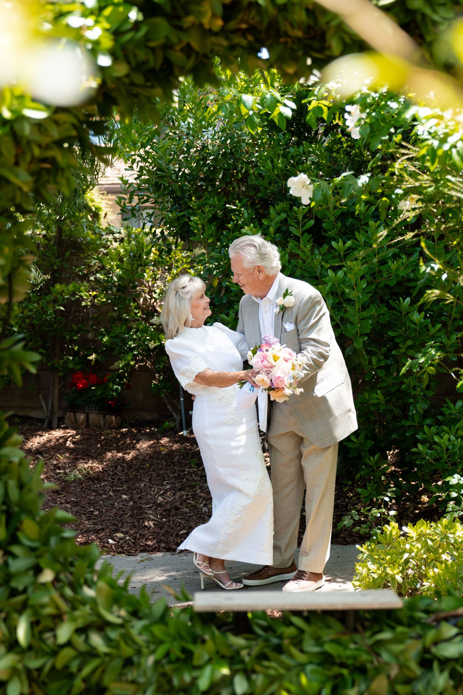 A couple sharing a romantic moment among lush garden greenery while holding a pink and white bouquet.