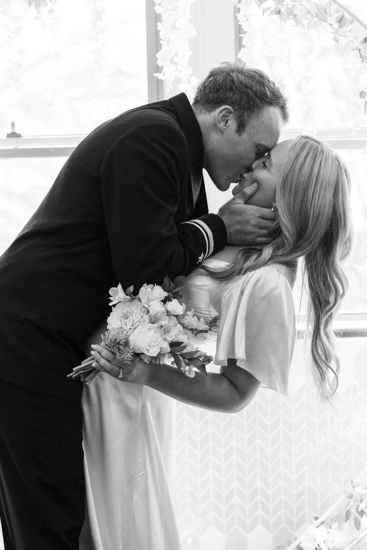 A romantic black and white photograph of a couple embracing at a wedding, with the bride holding a floral bouquet.