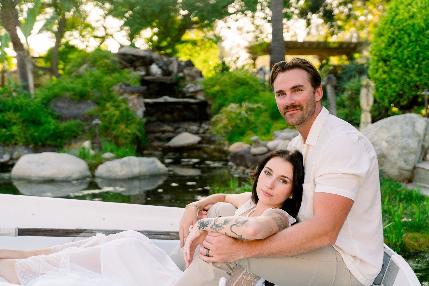 A couple relaxes outdoors near a garden waterfall feature on a sunny summer day.