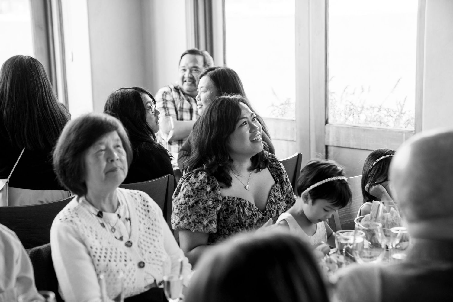 Wedding guests laugh and socialize at reception tables near bright windows.
