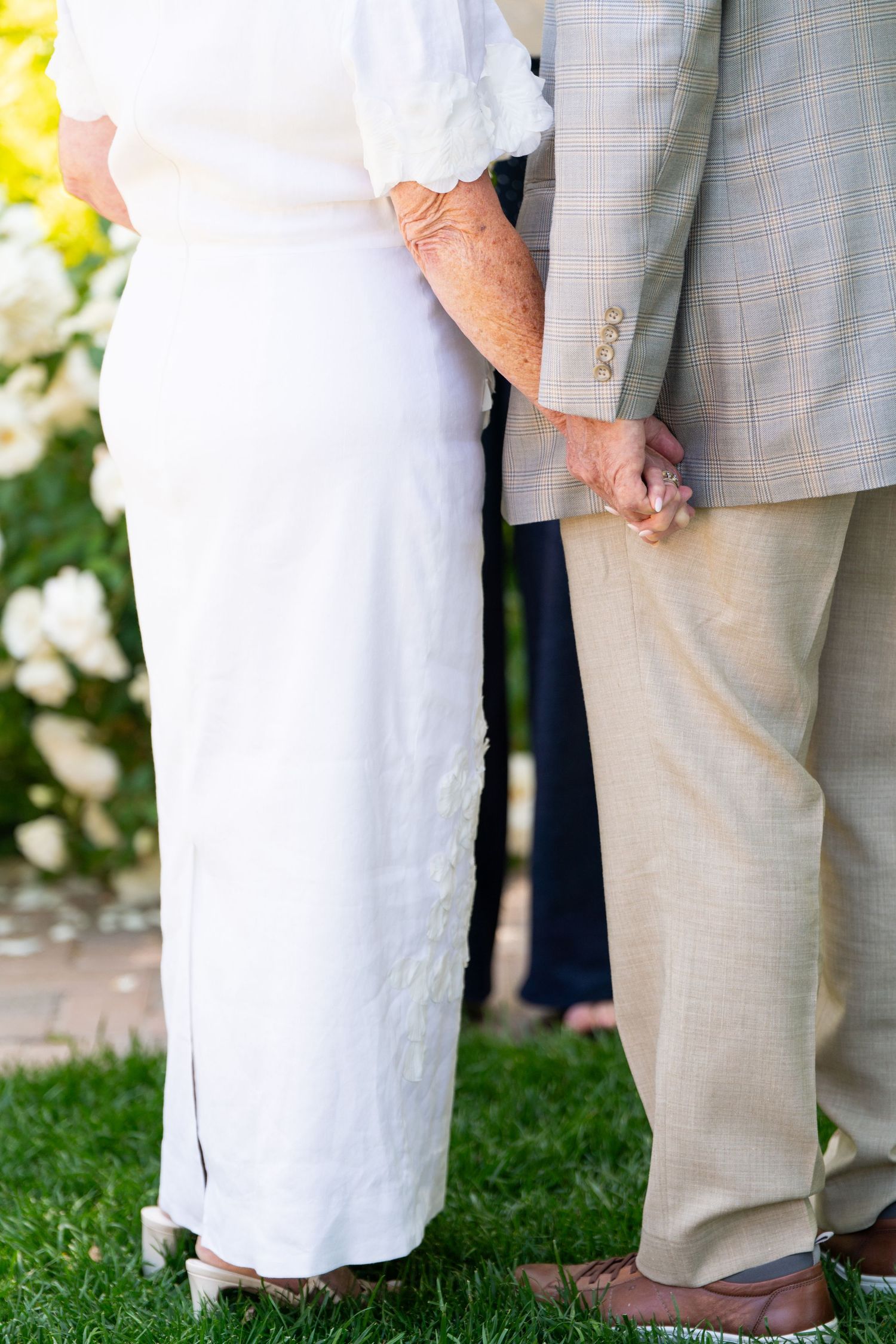 Close-up of a couple in white dress and grey suit holding hands during an outdoor ceremony.