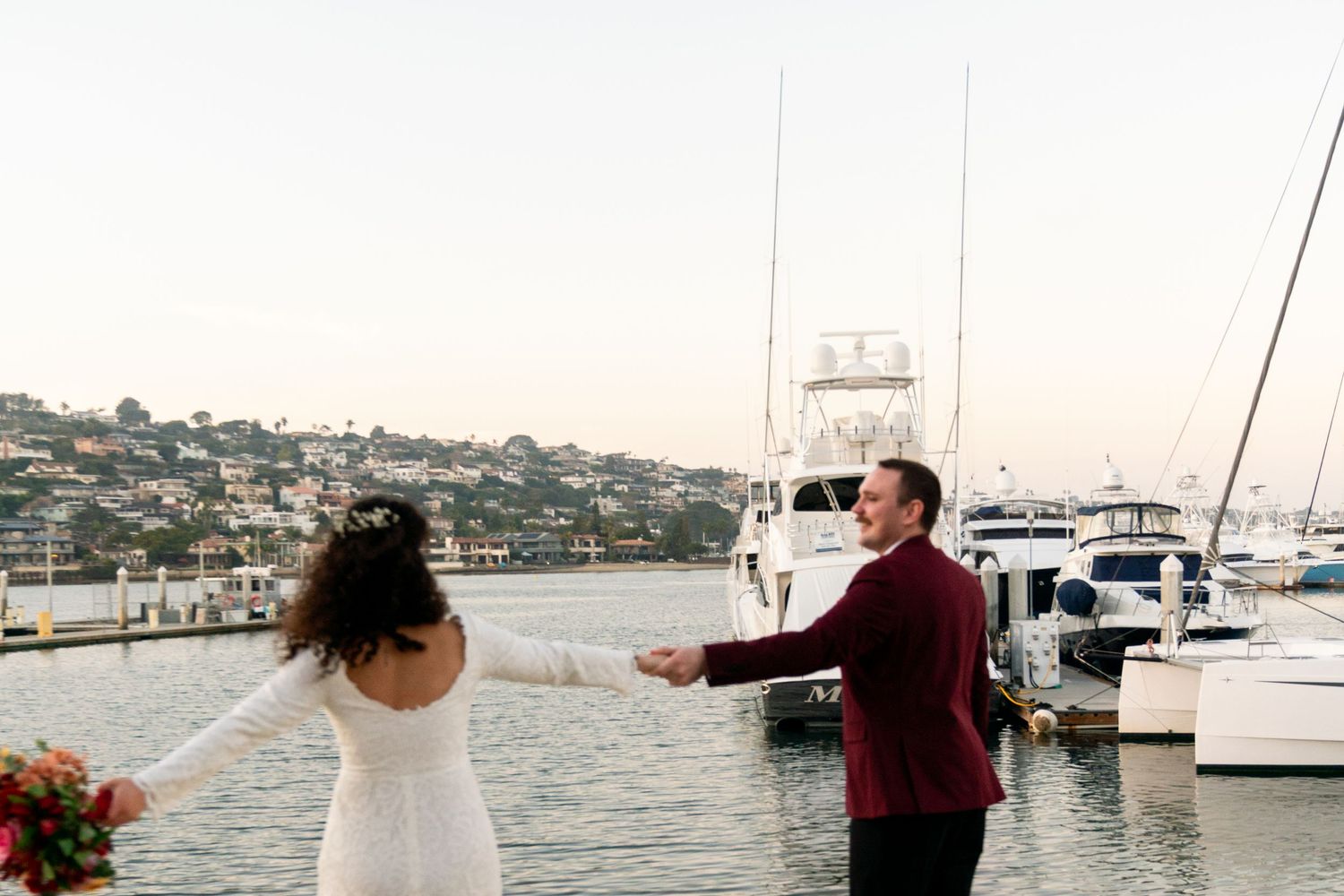 Couple dancing together on a marina dock with boats and hillside homes in the background at sunset.