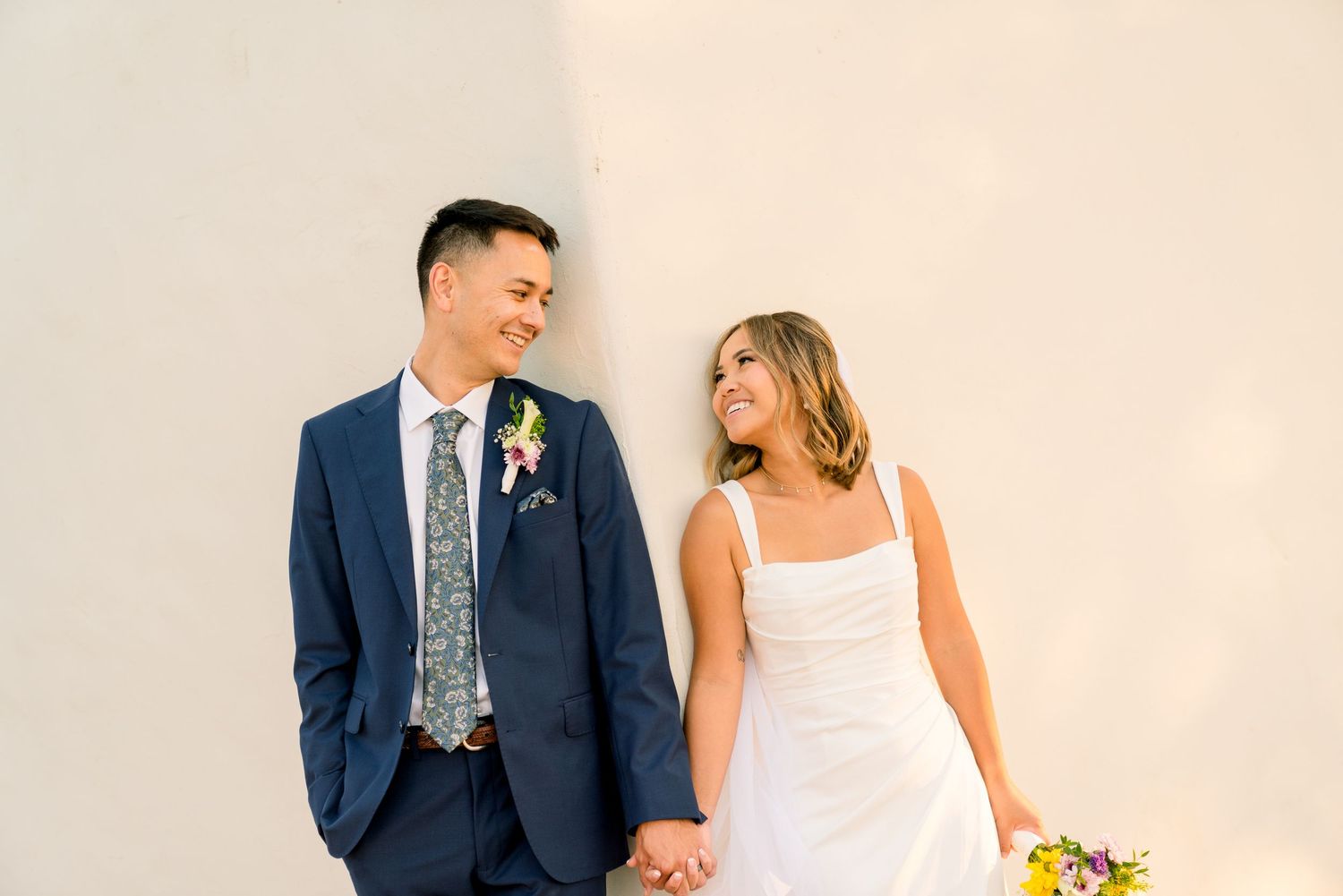A couple in wedding attire lean against a beige wall while holding hands and smiling at each other.