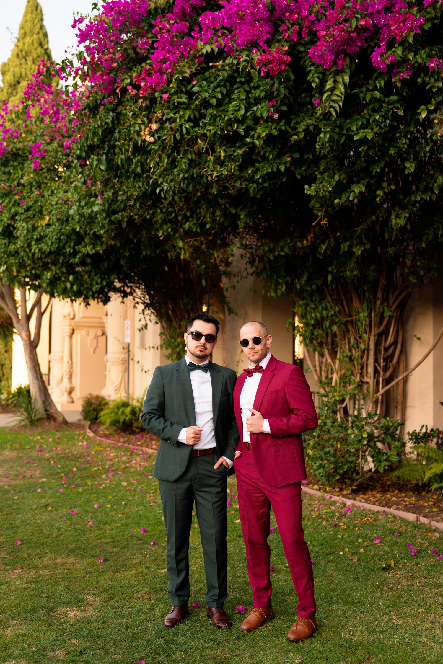 Two people in formal attire posing in front of vibrant pink bougainvillea flowers and garden architecture.