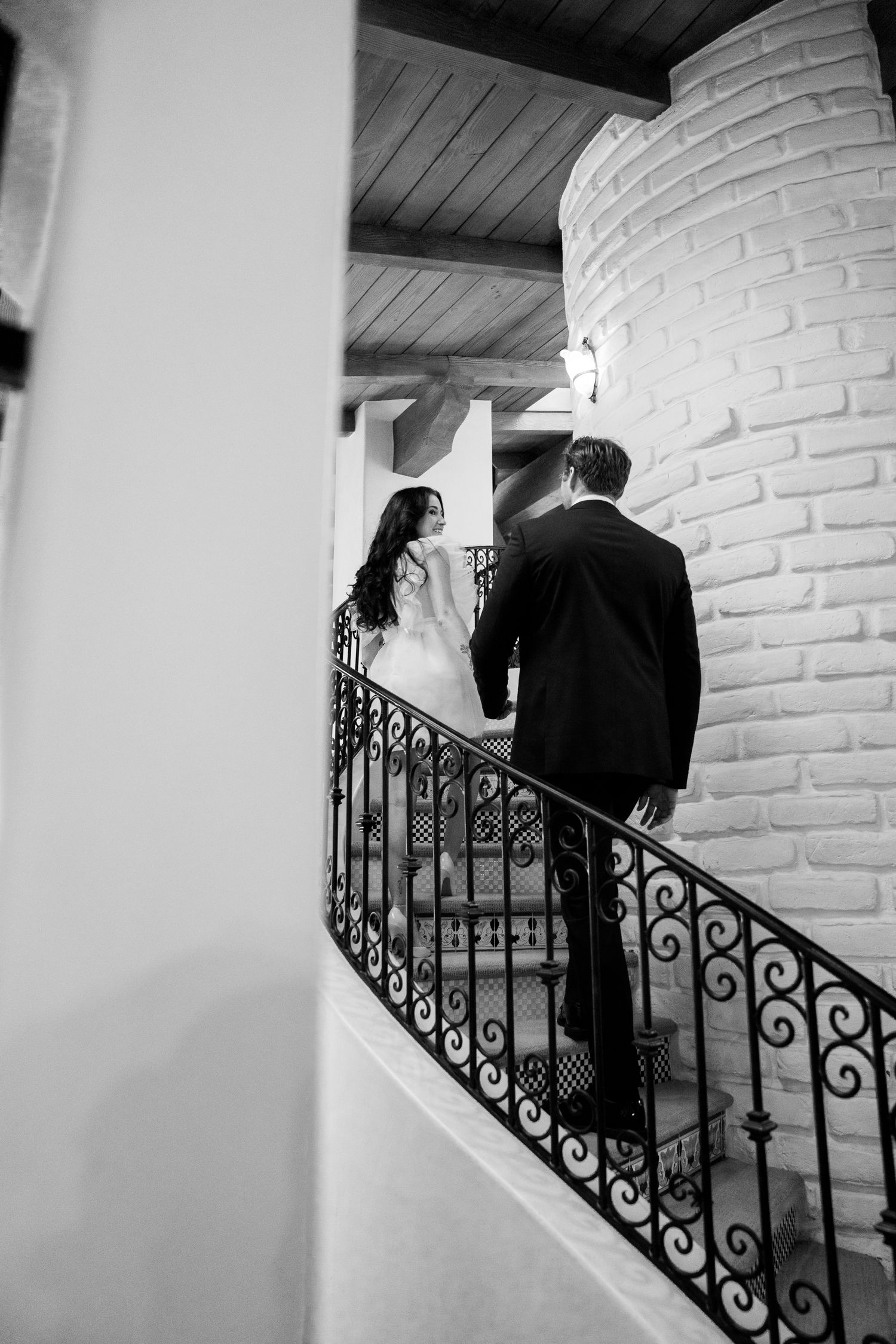 Black and white image of couple ascending ornate staircase with wrought iron railing.
