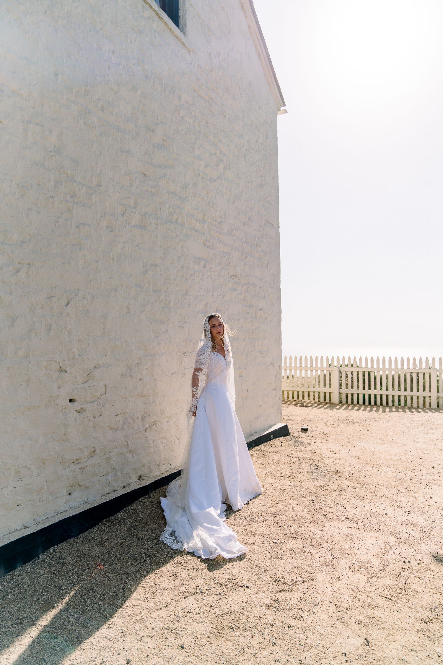 A bride in a flowing white wedding dress stands against a white building wall near a picket fence on a sunny day.