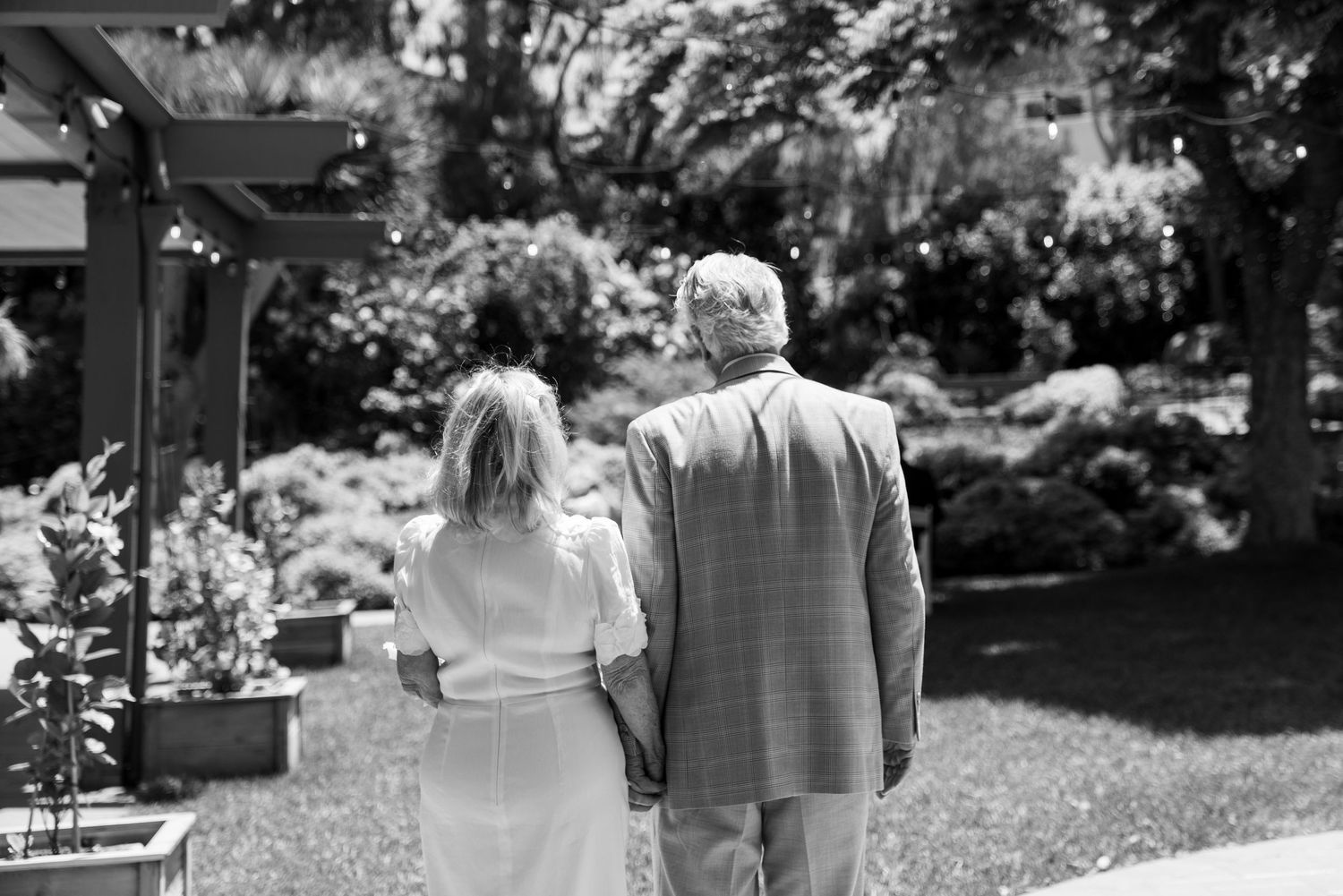 A couple stands together in a garden setting, photographed from behind in black and white.