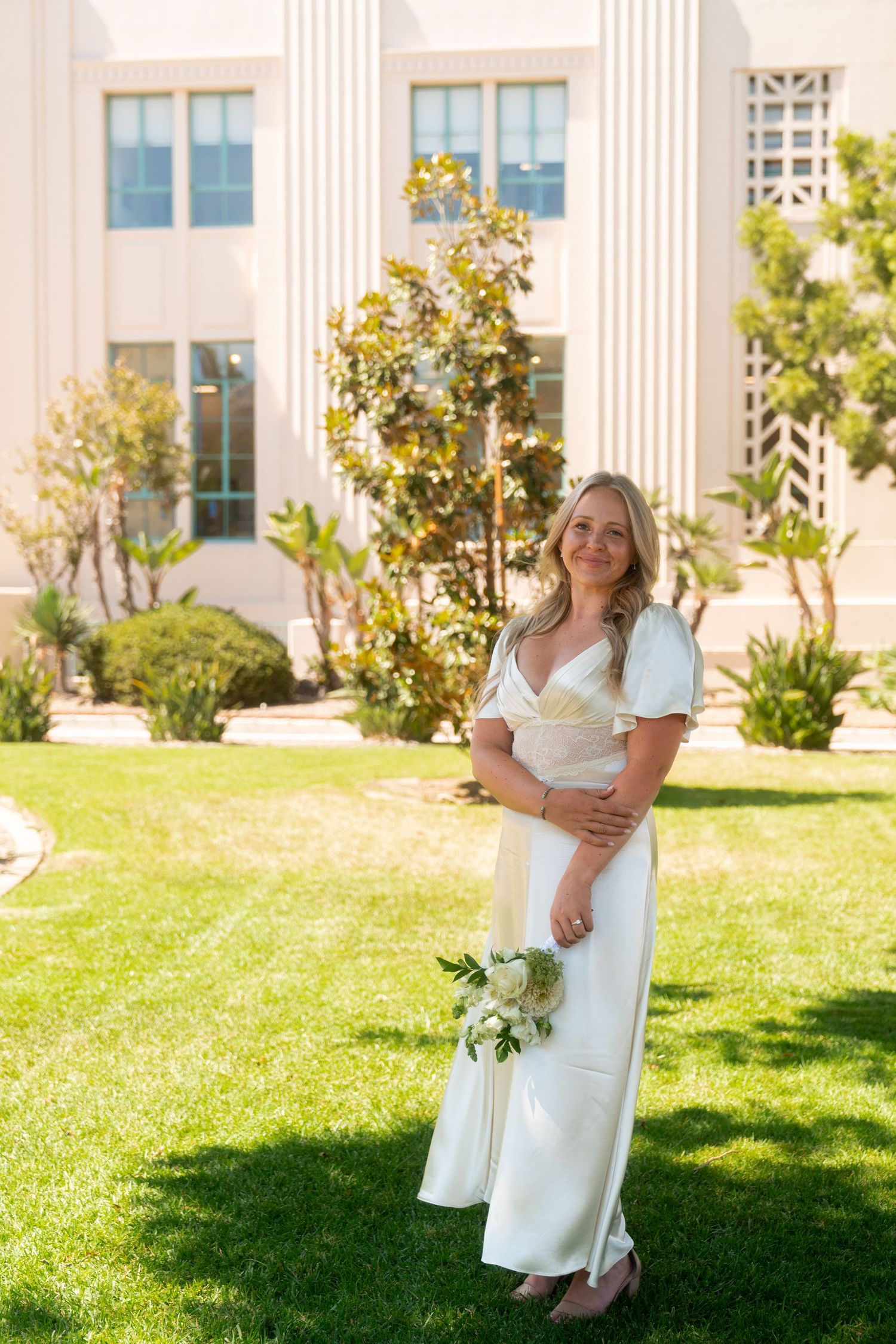 A person in a white dress stands on manicured lawn in front of a classical building with columns on a sunny day.