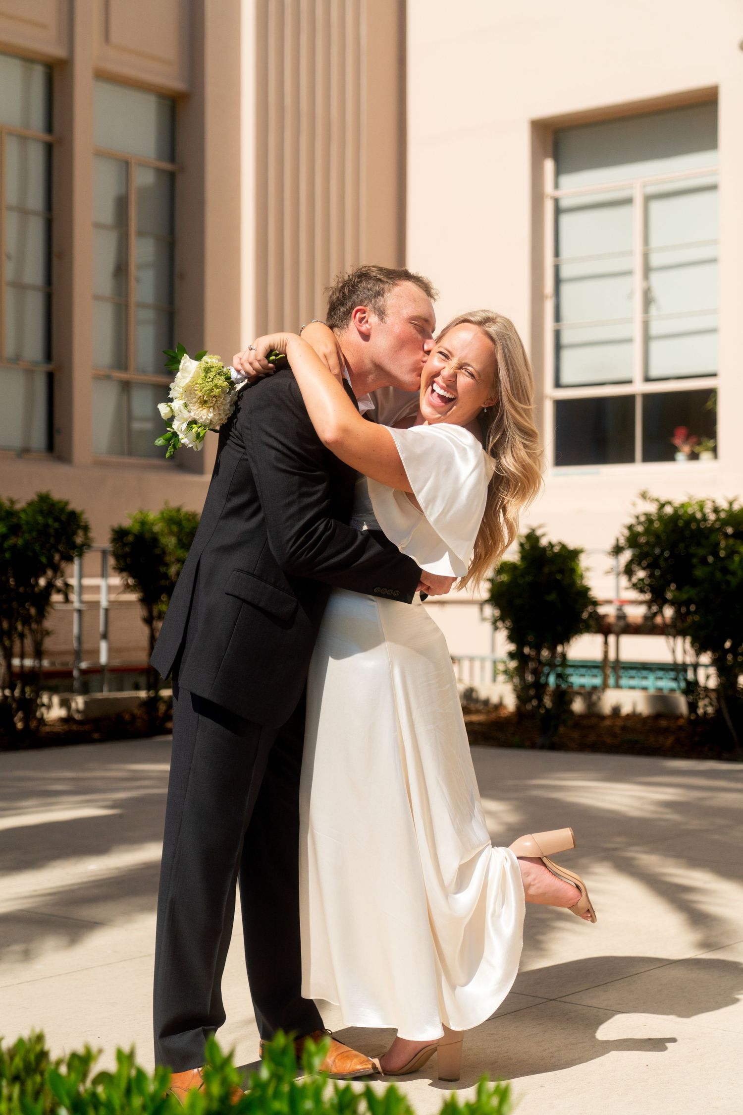 A couple in wedding attire shares romantic moments during a sunset photo session outside a building.
