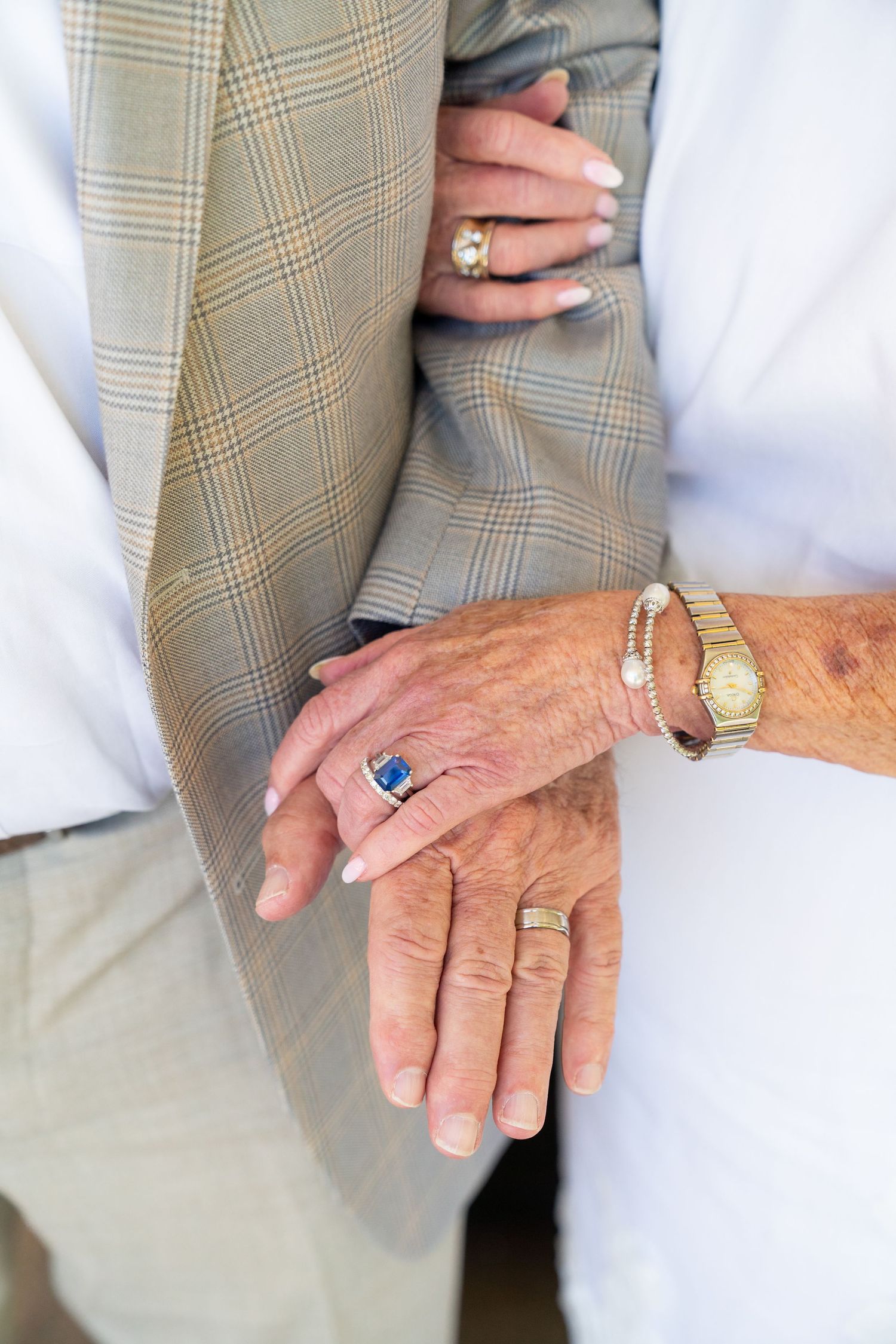 Close-up shot of hands wearing wedding rings, a blue gemstone ring, and a gold watch against a plaid suit jacket.