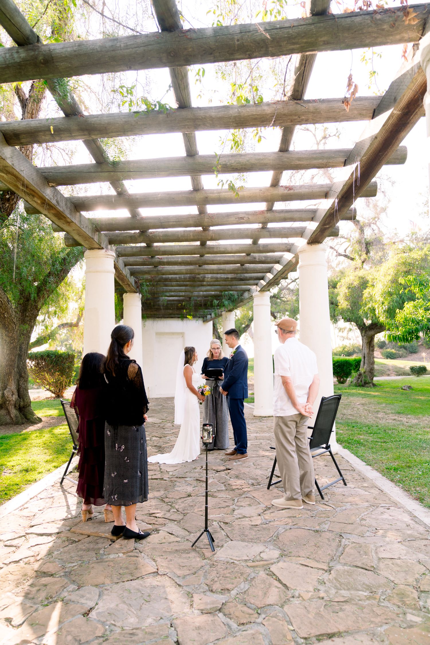 Small outdoor ceremony taking place under rustic wooden pergola on stone patio surrounded by garden scenery.