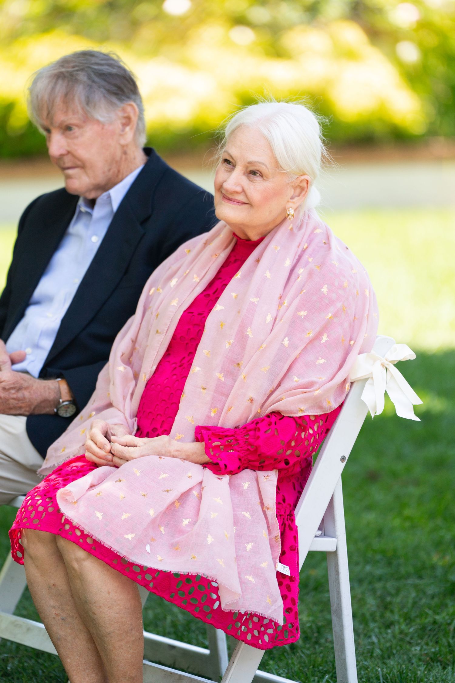 Senior couple sitting outdoors, with woman wearing pink shawl and red dress at sunny garden event.