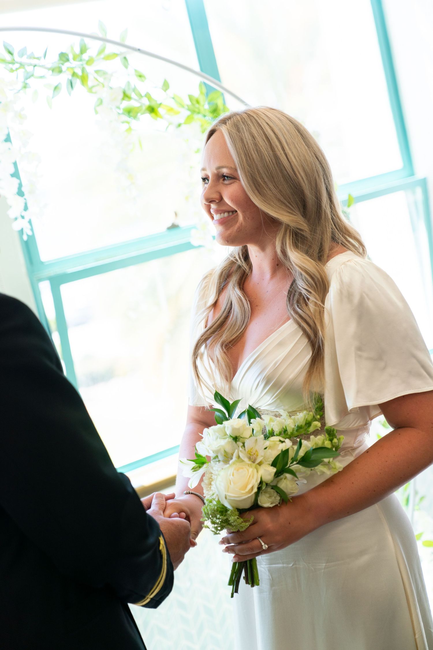 Bride in white wedding dress with flutter sleeves holds elegant white and cream floral bouquet at indoor ceremony.