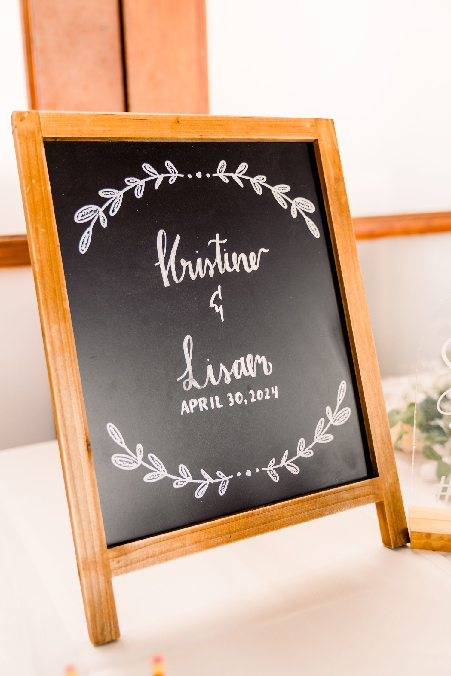 Rustic wooden-framed chalkboard sign displaying couple names and wedding date with decorative leaf wreath designs.