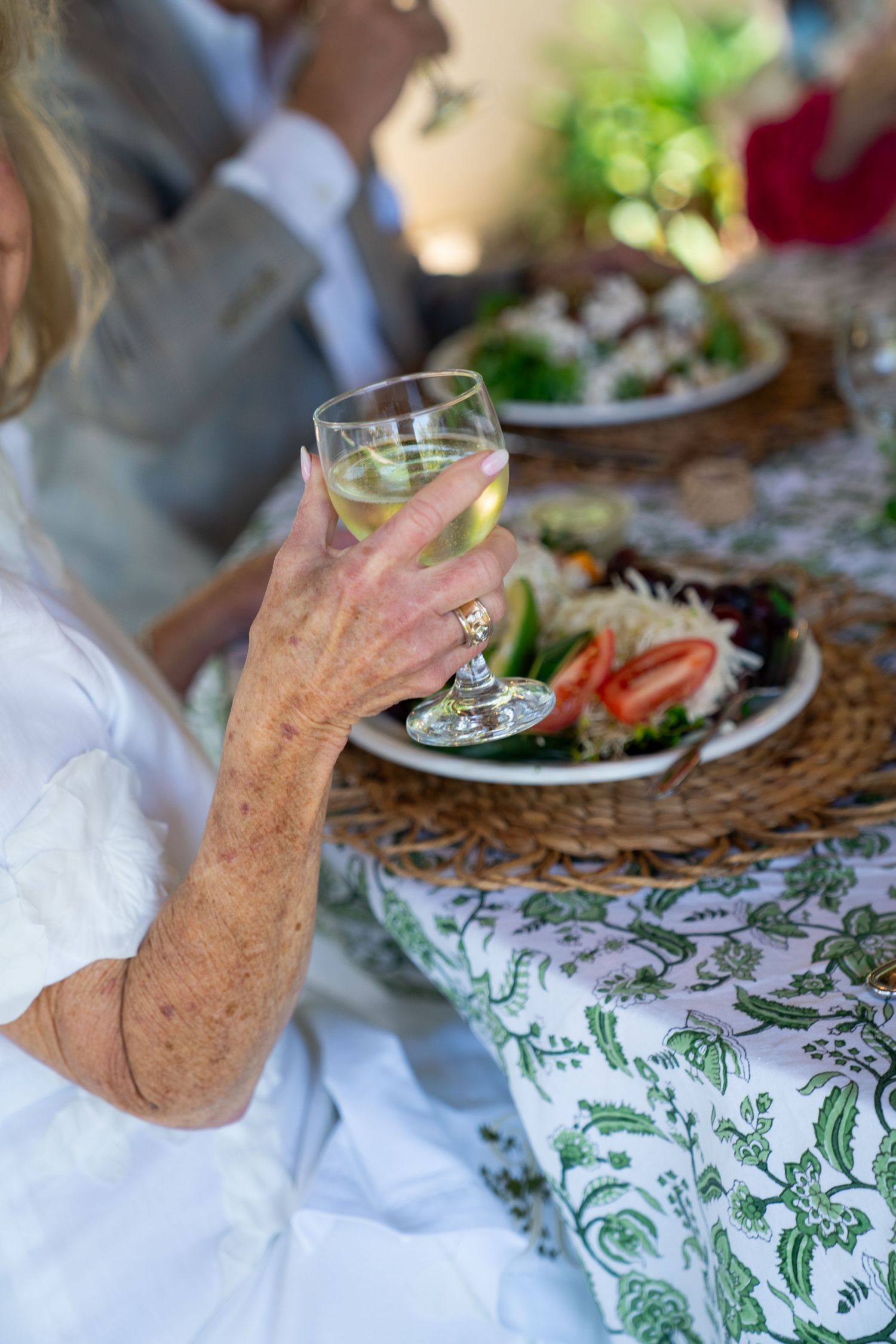 A hand holding a wine glass at an outdoor dining table with elegant place settings and patterned tablecloth.