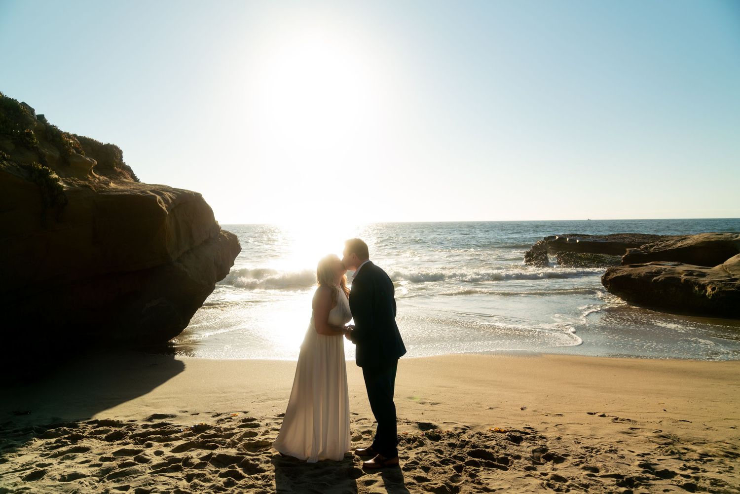 Silhouetted couple embracing on sandy beach at sunset with rocky cliffs and ocean waves in background.