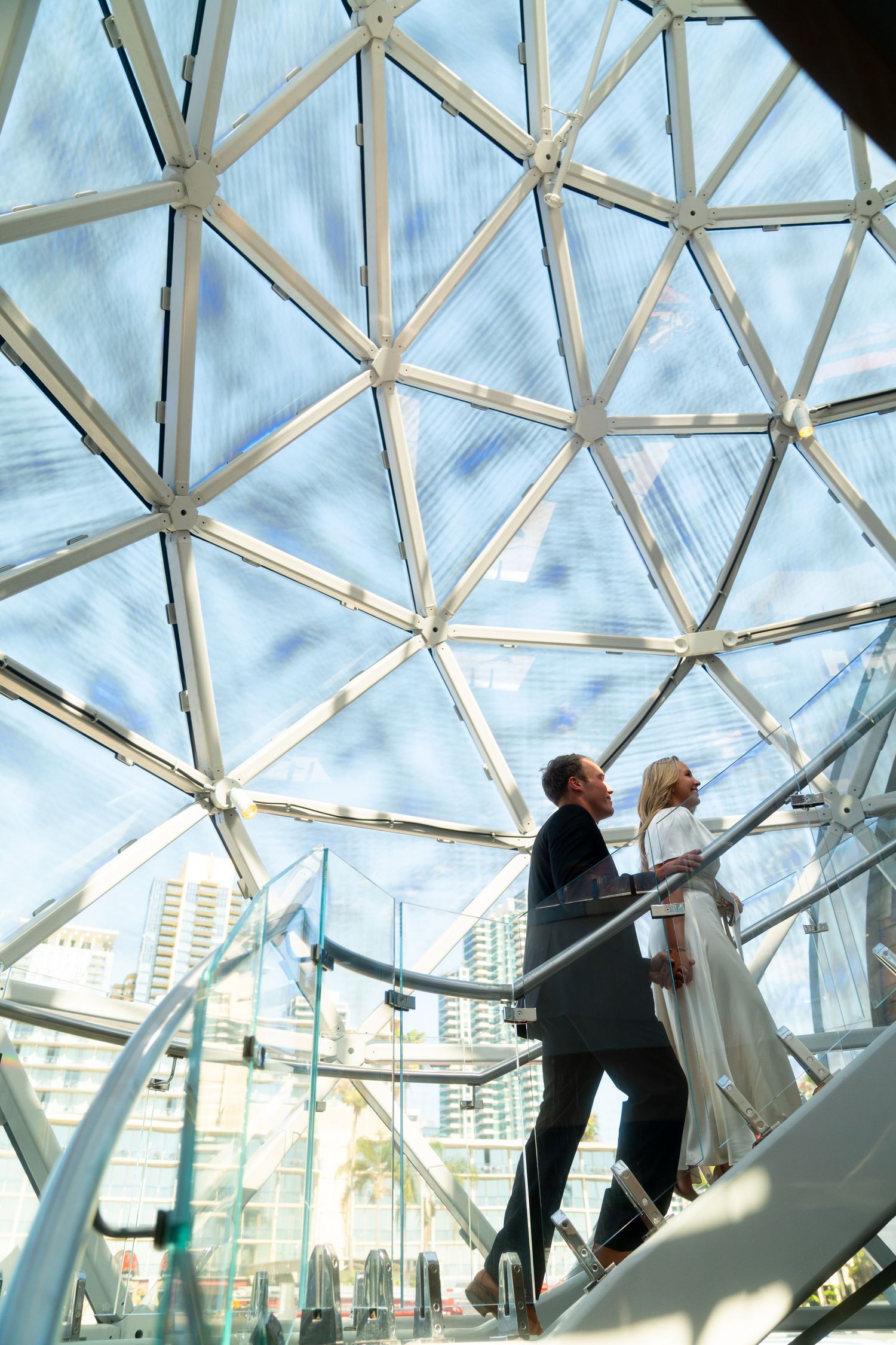 A couple poses beneath a geometric glass dome ceiling in a modern architectural space, with city buildings visible outside.