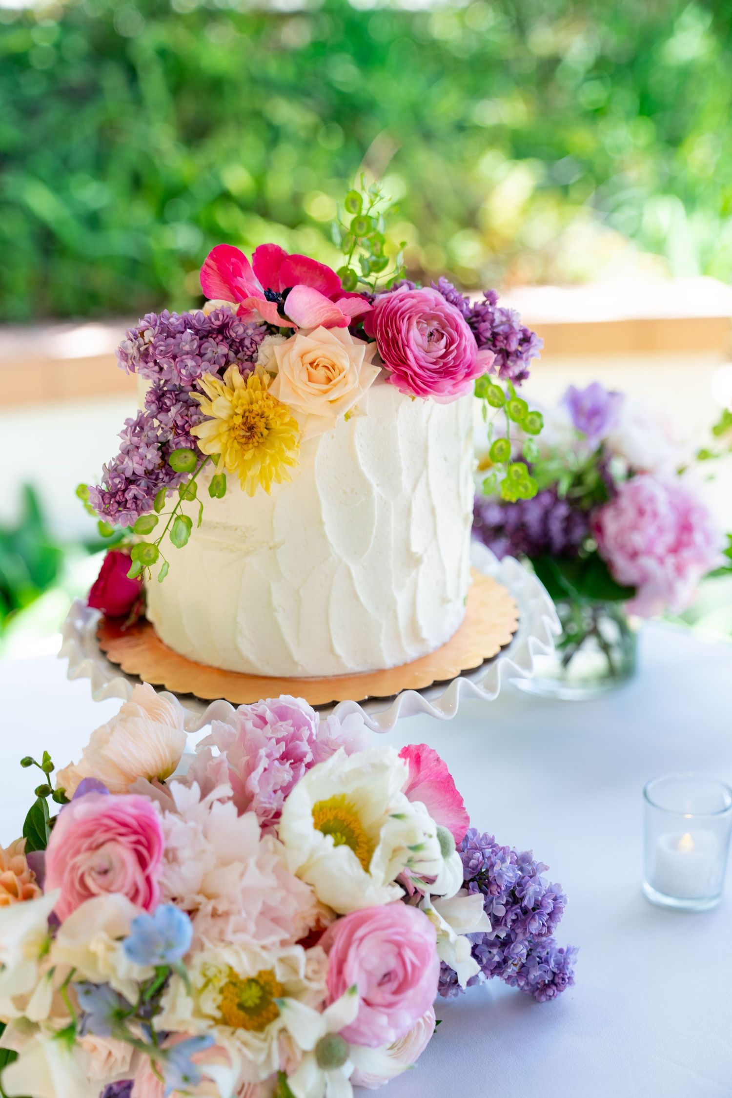 White wedding cake decorated with colorful fresh flowers including roses, peonies and lilacs on table setting.