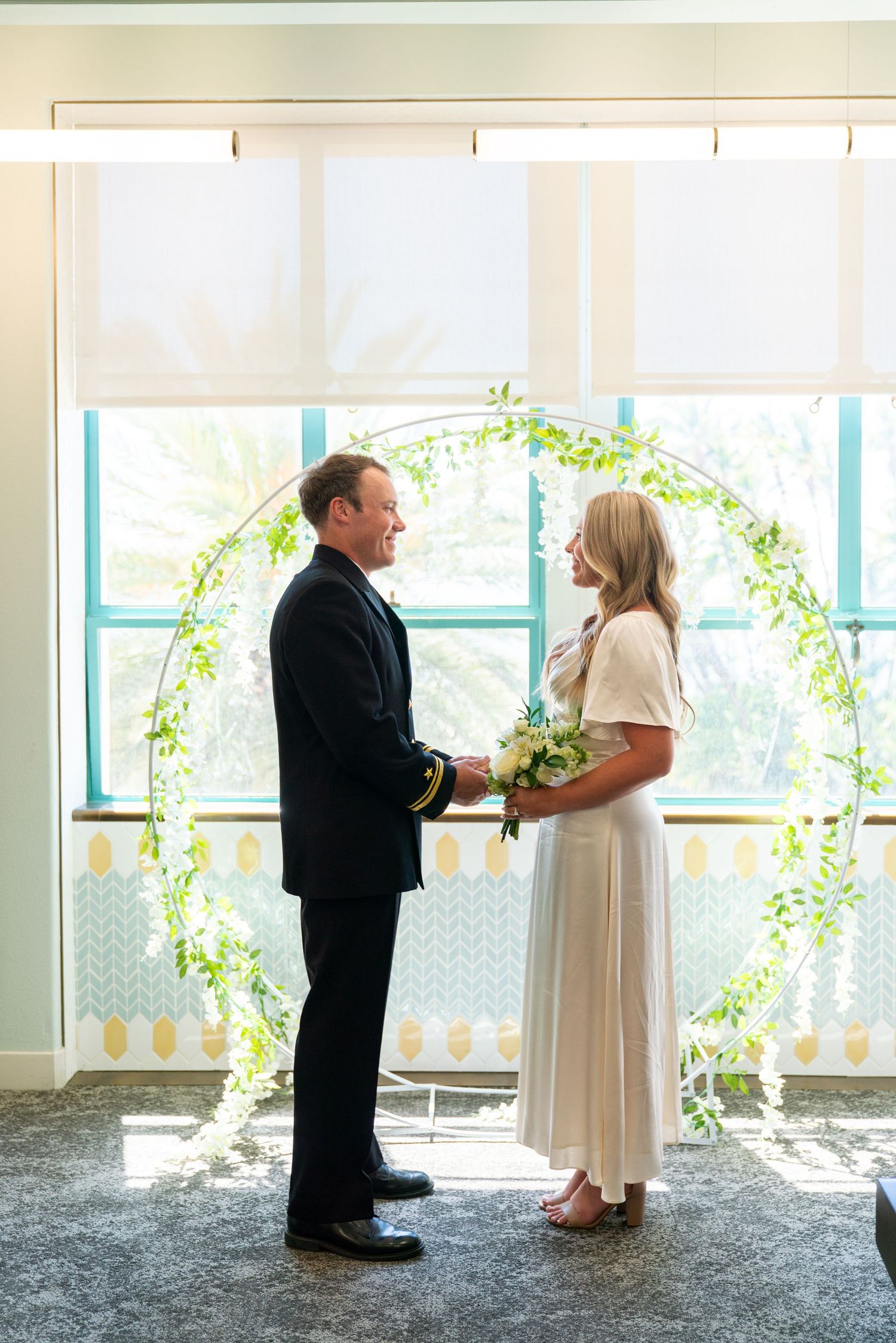 A couple stands together under a floral arch during an intimate wedding ceremony in a light-filled indoor space.