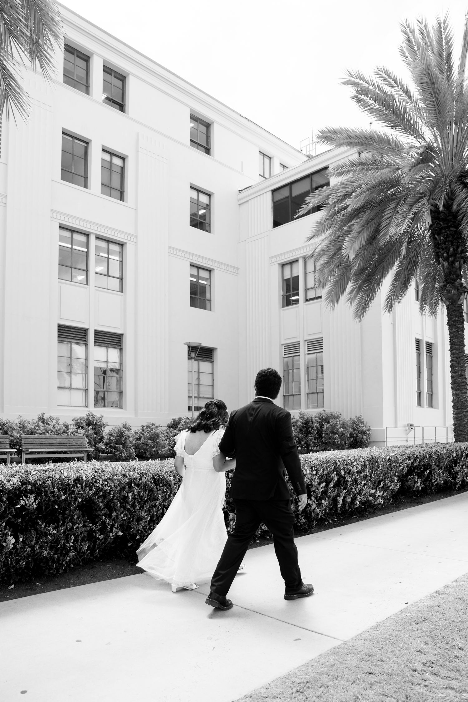 Black and white photo of couple walking along path beside white building with palm tree.