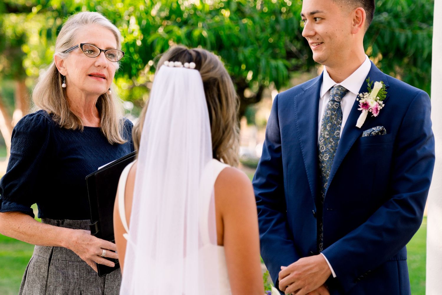 An outdoor wedding ceremony takes place in a garden setting with guests and wedding party members present.