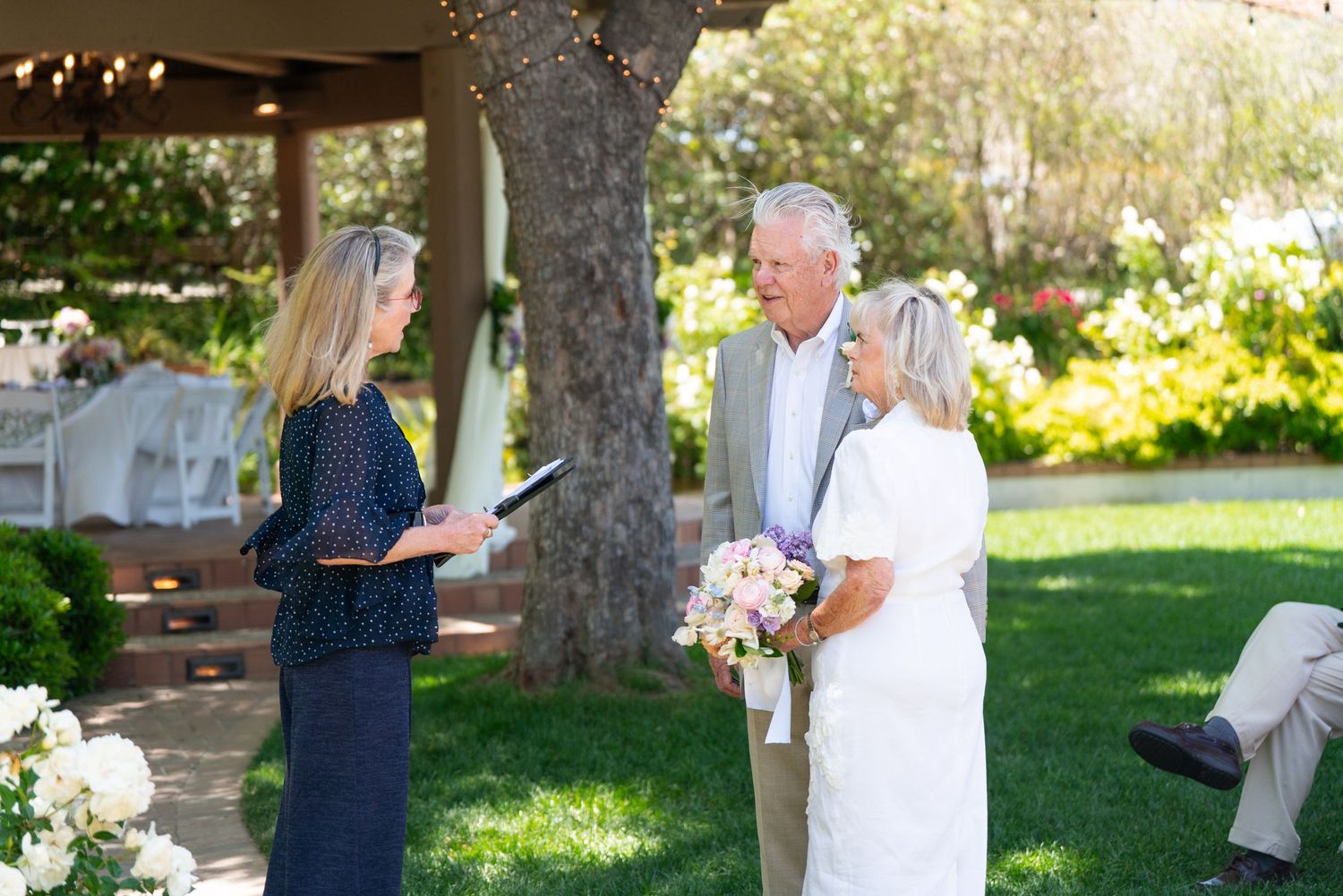 An outdoor ceremony takes place in a garden setting with people gathered under a tree on a sunny day.