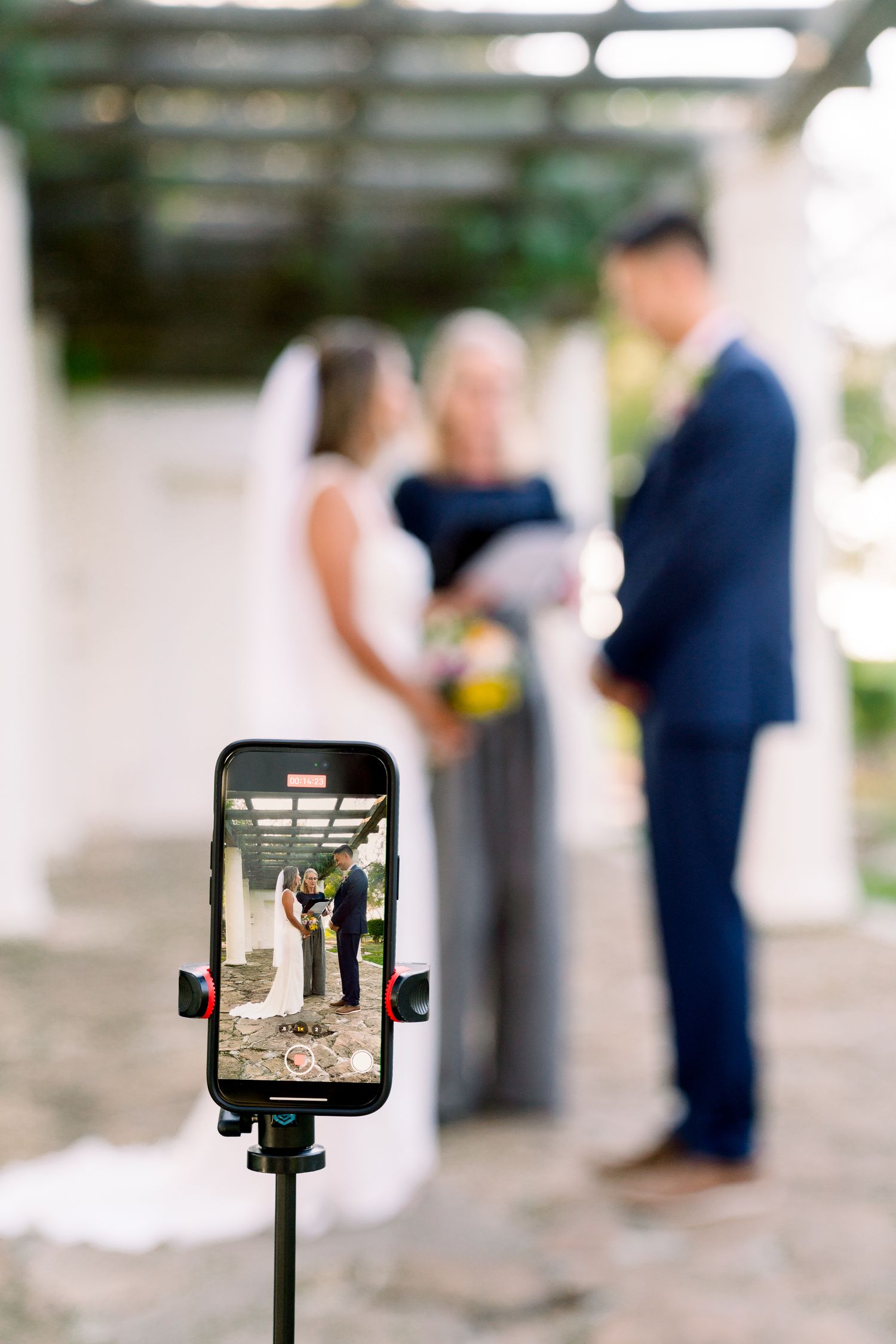 A wedding ceremony being livestreamed on a mobile phone mounted on a tripod in the foreground.