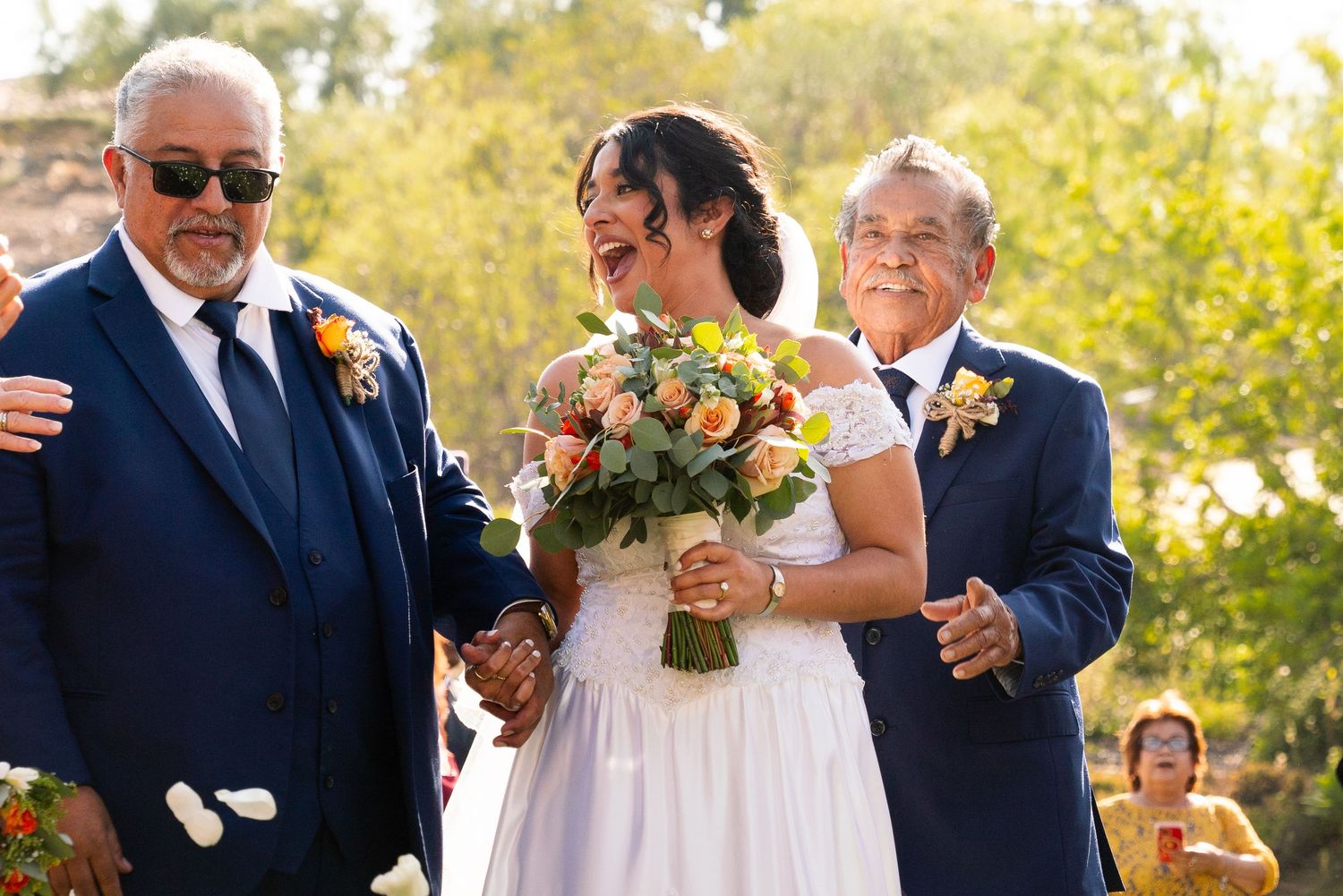A smiling bride in a white dress holds a peach and orange bouquet while being escorted down an outdoor aisle.