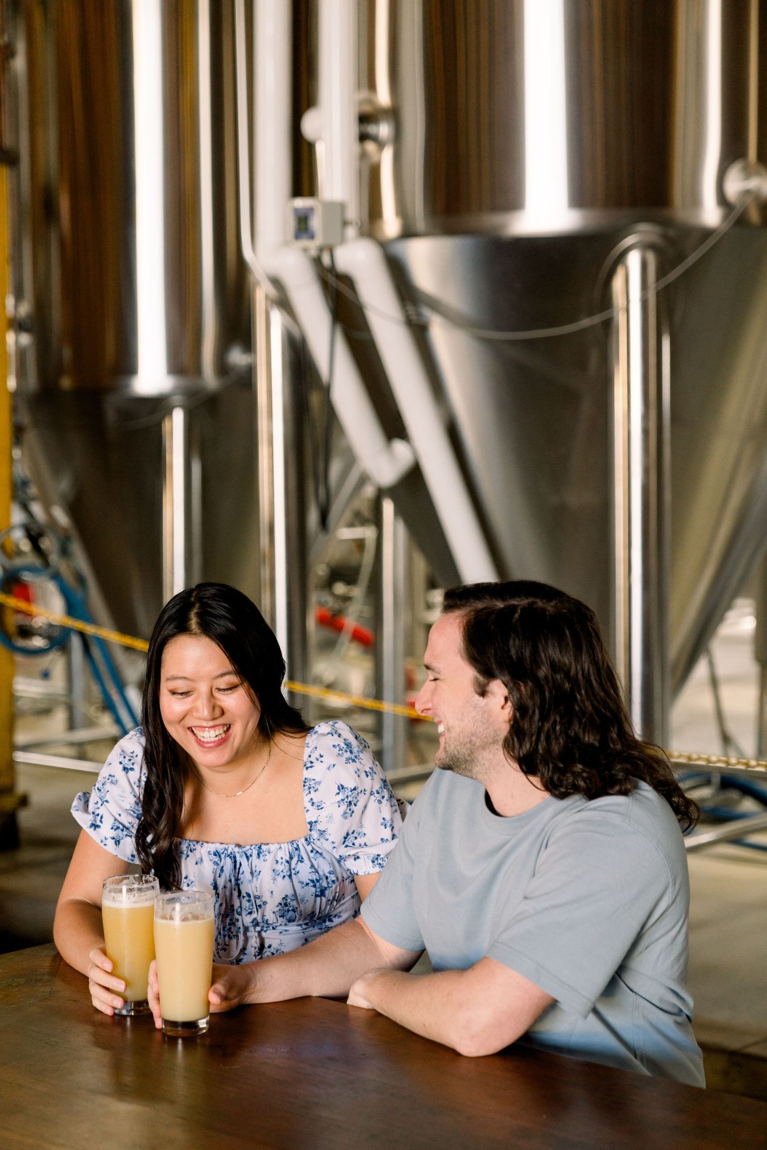 Couple enjoying drinks together at brewery with steel fermentation tanks in background.