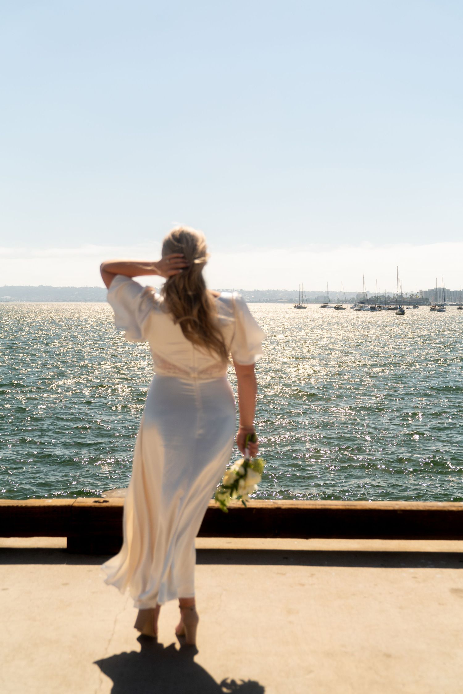 Woman in flowing white dress stands at waterfront overlooking marina on sunny day with sparkling water and boats in distance.