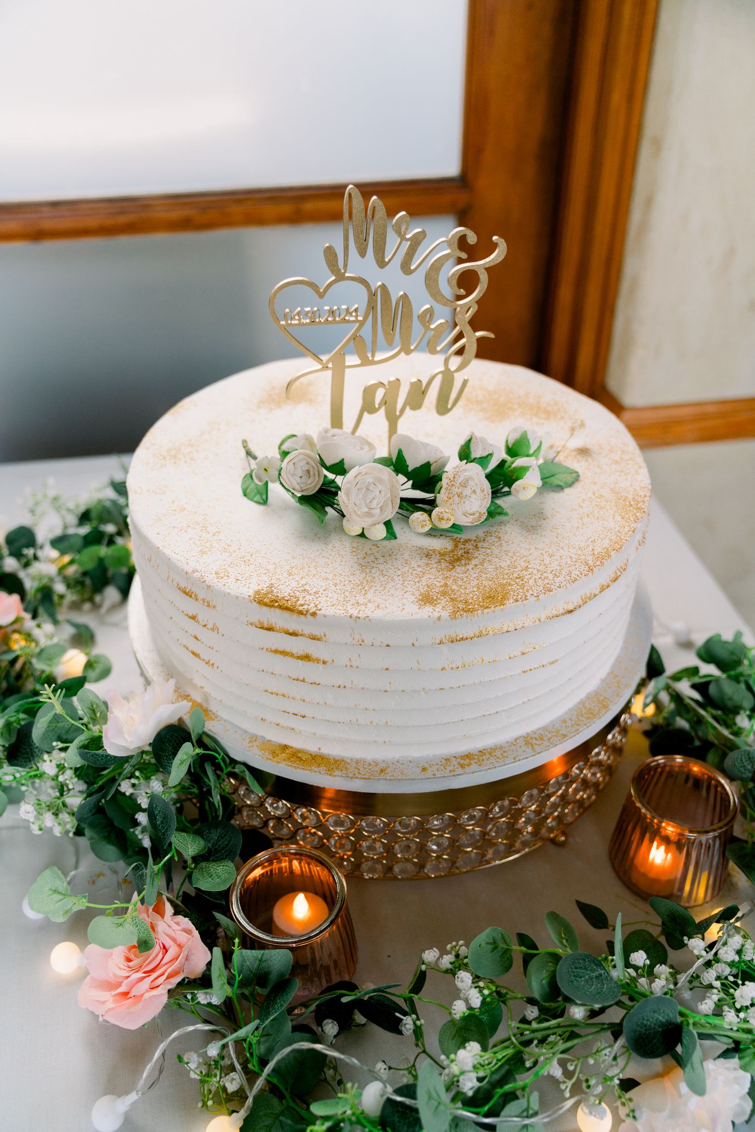 Elegant white wedding cake with gold accents and floral decorations on a crystal stand surrounded by candles and greenery.