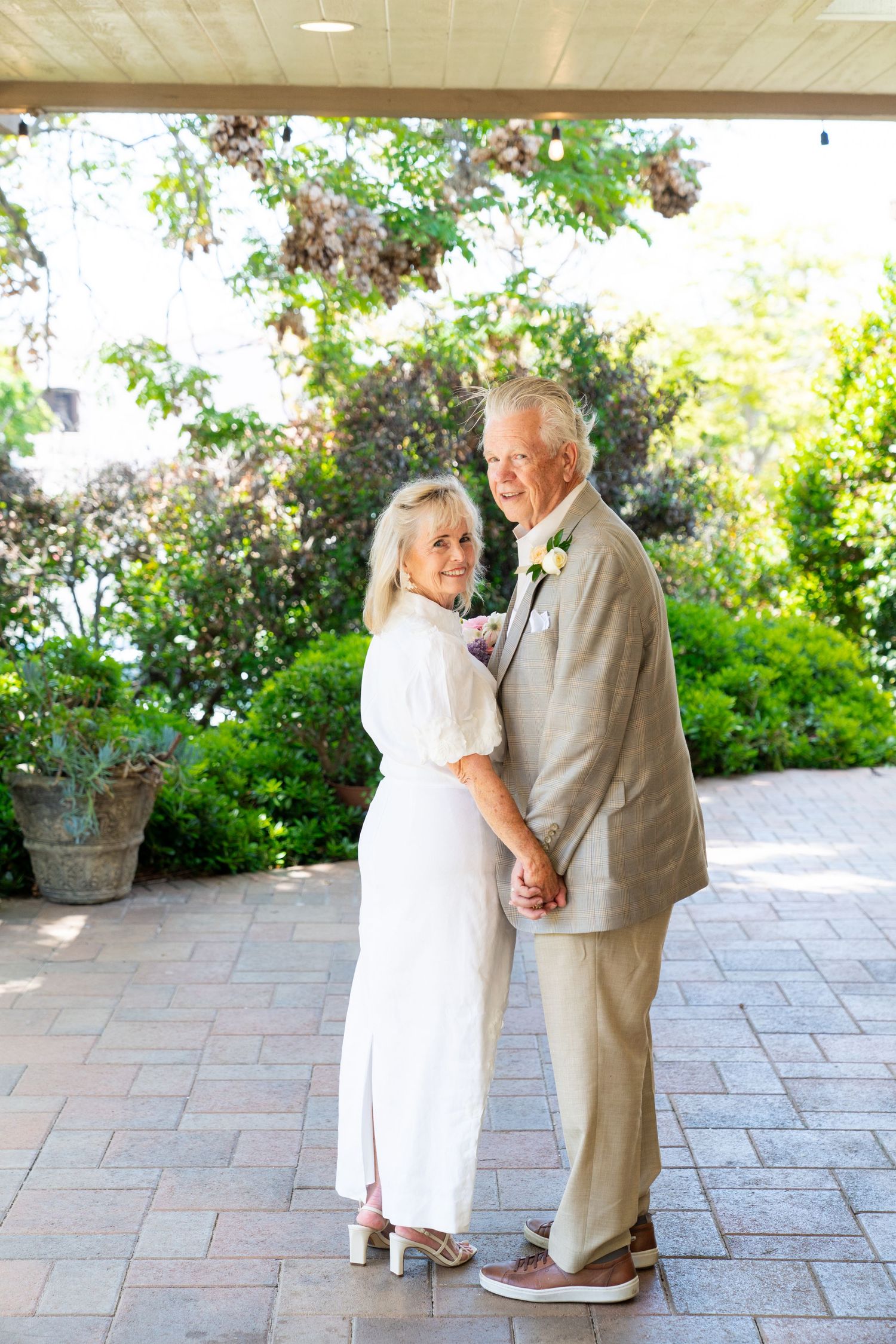 A couple in elegant white and beige attire holding hands on a patio with greenery and garden decor in the background.