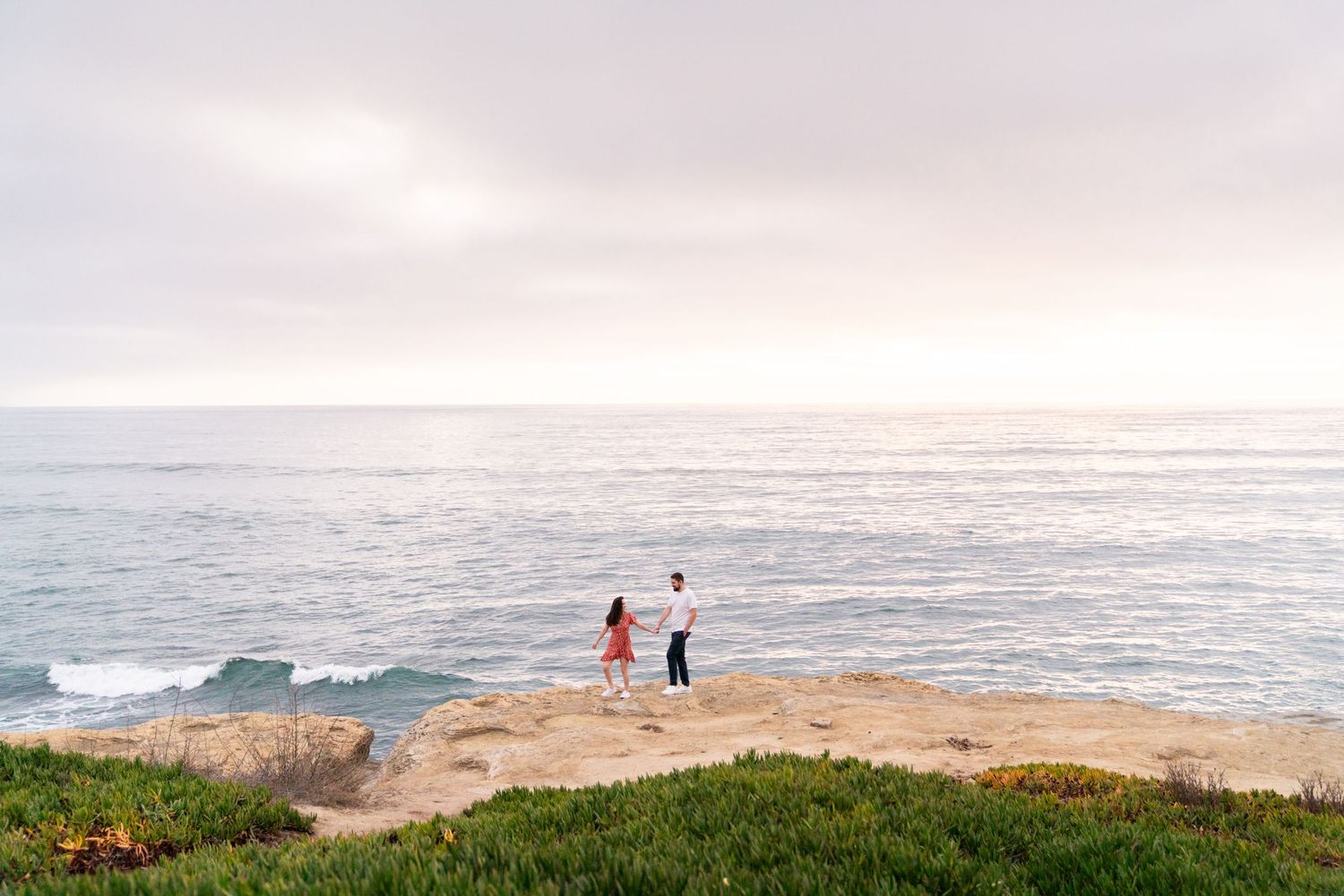 Two people stand on a coastal cliff edge against a soft cloudy sky and vast ocean view.