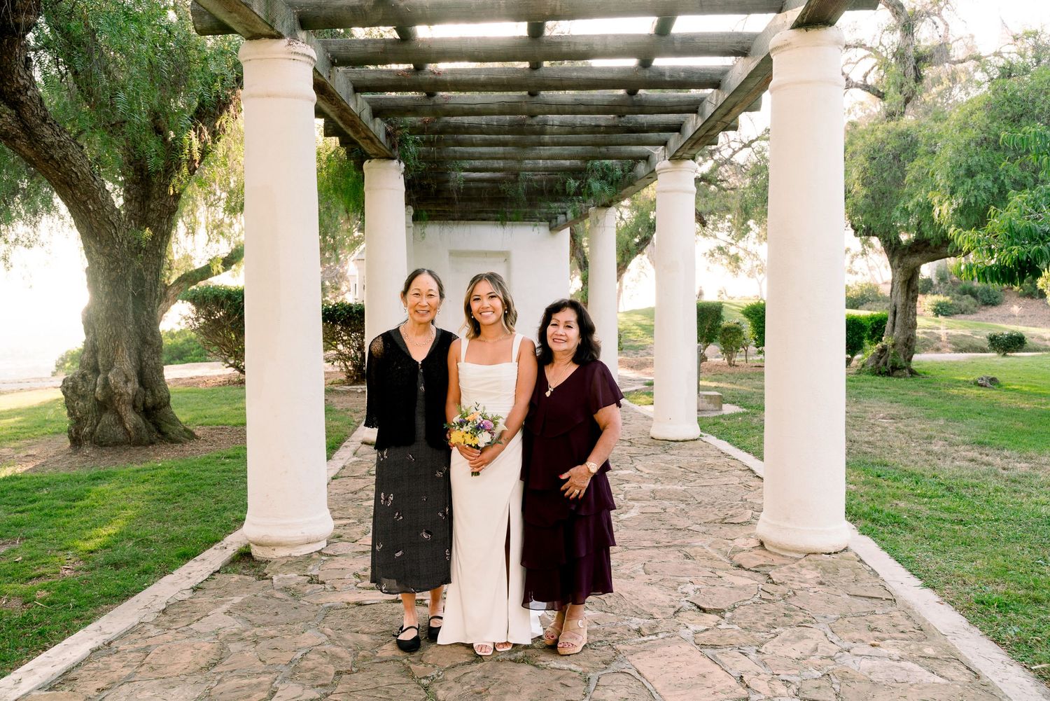 Three people stand under an elegant covered walkway with white columns and pergola roof in an outdoor garden setting.