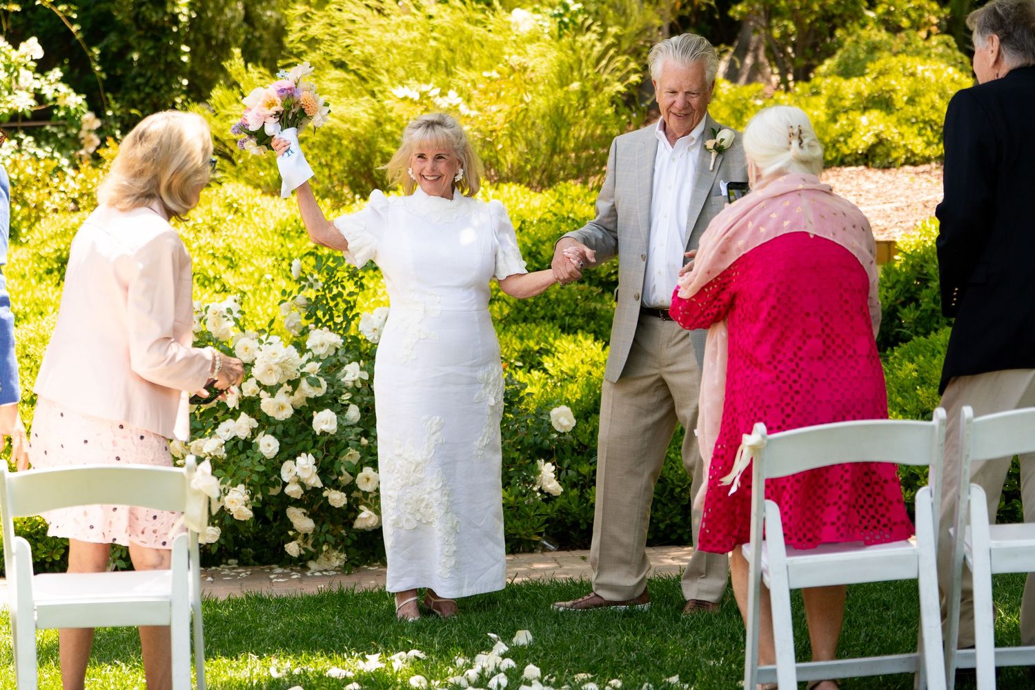 A group of people gather for a celebration in a sunny garden with blooming white flowers and lush greenery.