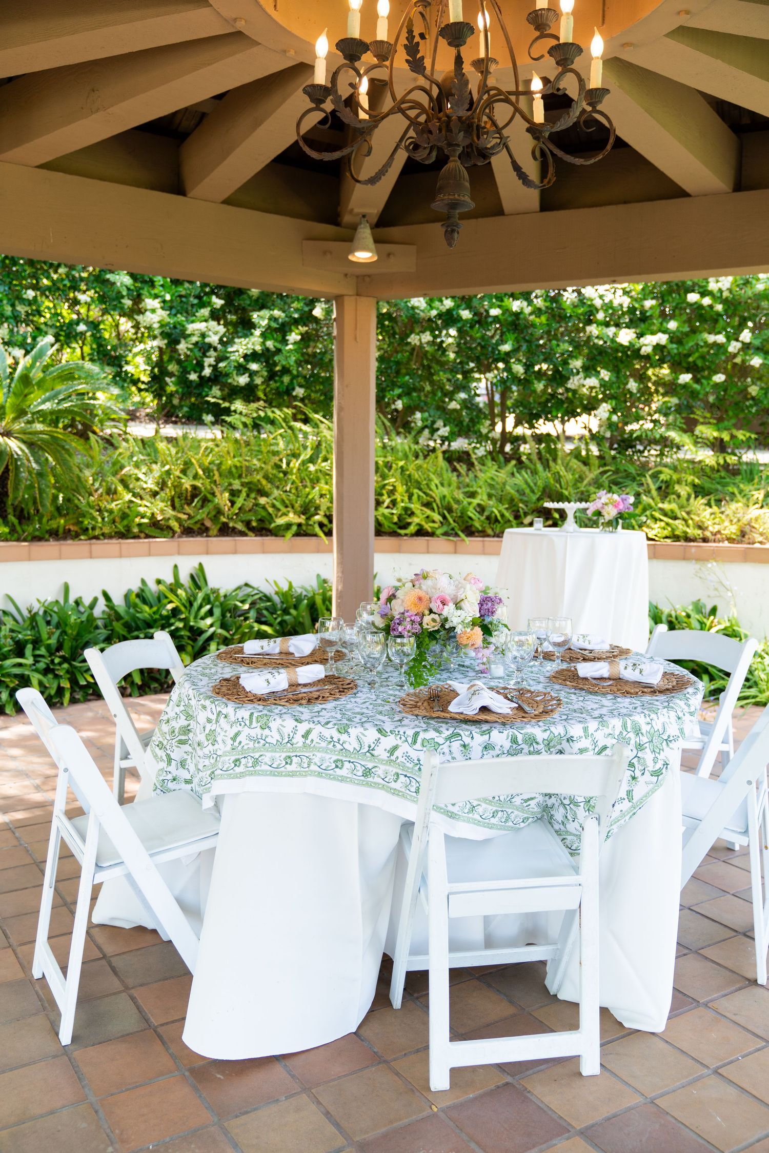 Elegant outdoor dining table set with white chairs and floral tablecloth under a gazebo with chandelier.