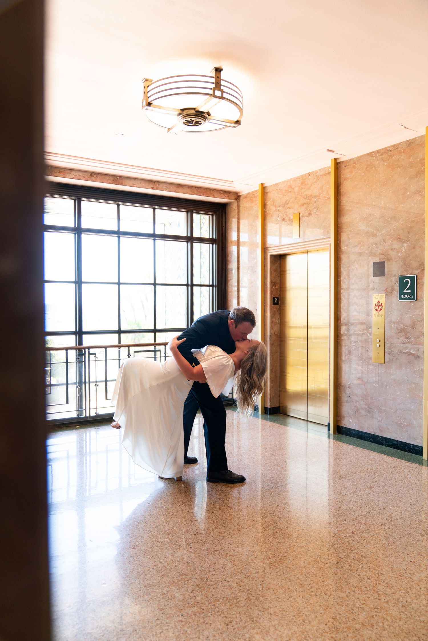 A romantic dance dip pose in a bright hallway with elevators and a decorative ceiling light.