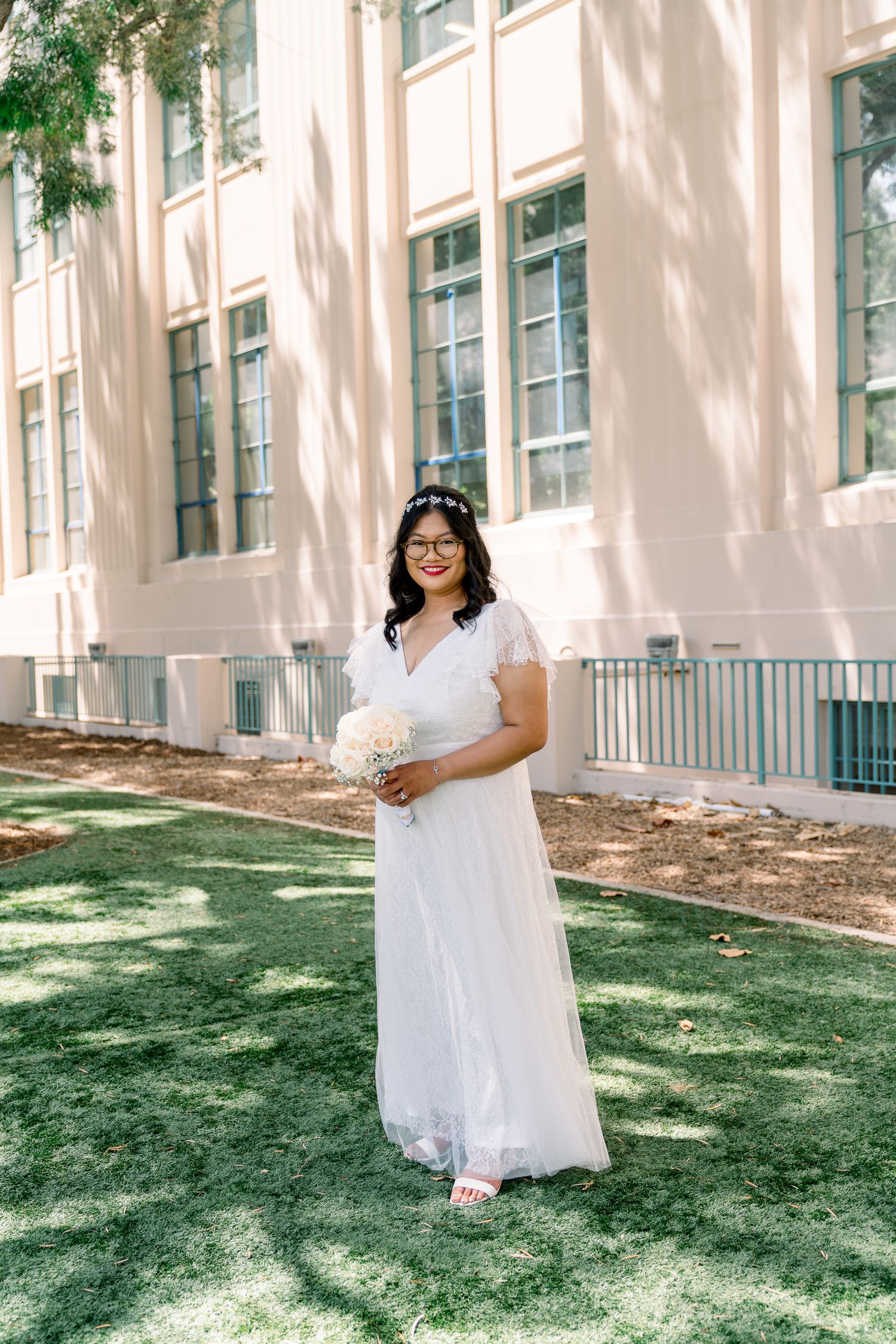A bride in a white lace dress and glasses poses outdoors in front of a pink building on a sunny day.