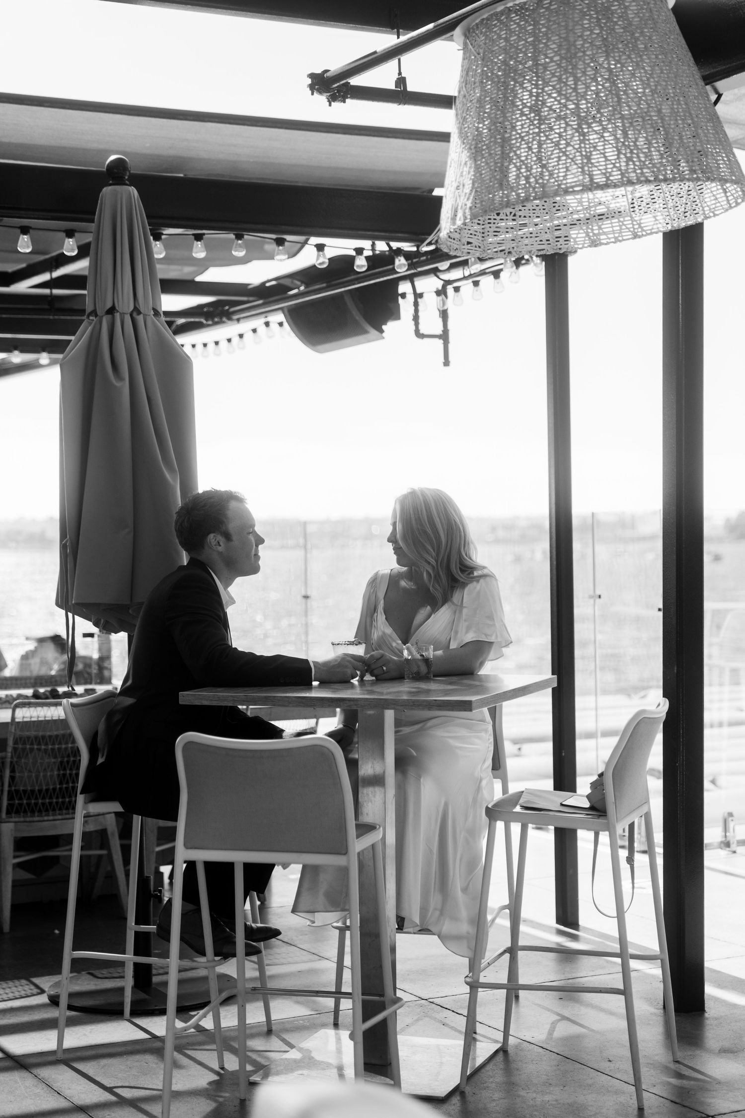 Two people sit at a high top table by large windows in a restaurant, having an intimate conversation in black and white.