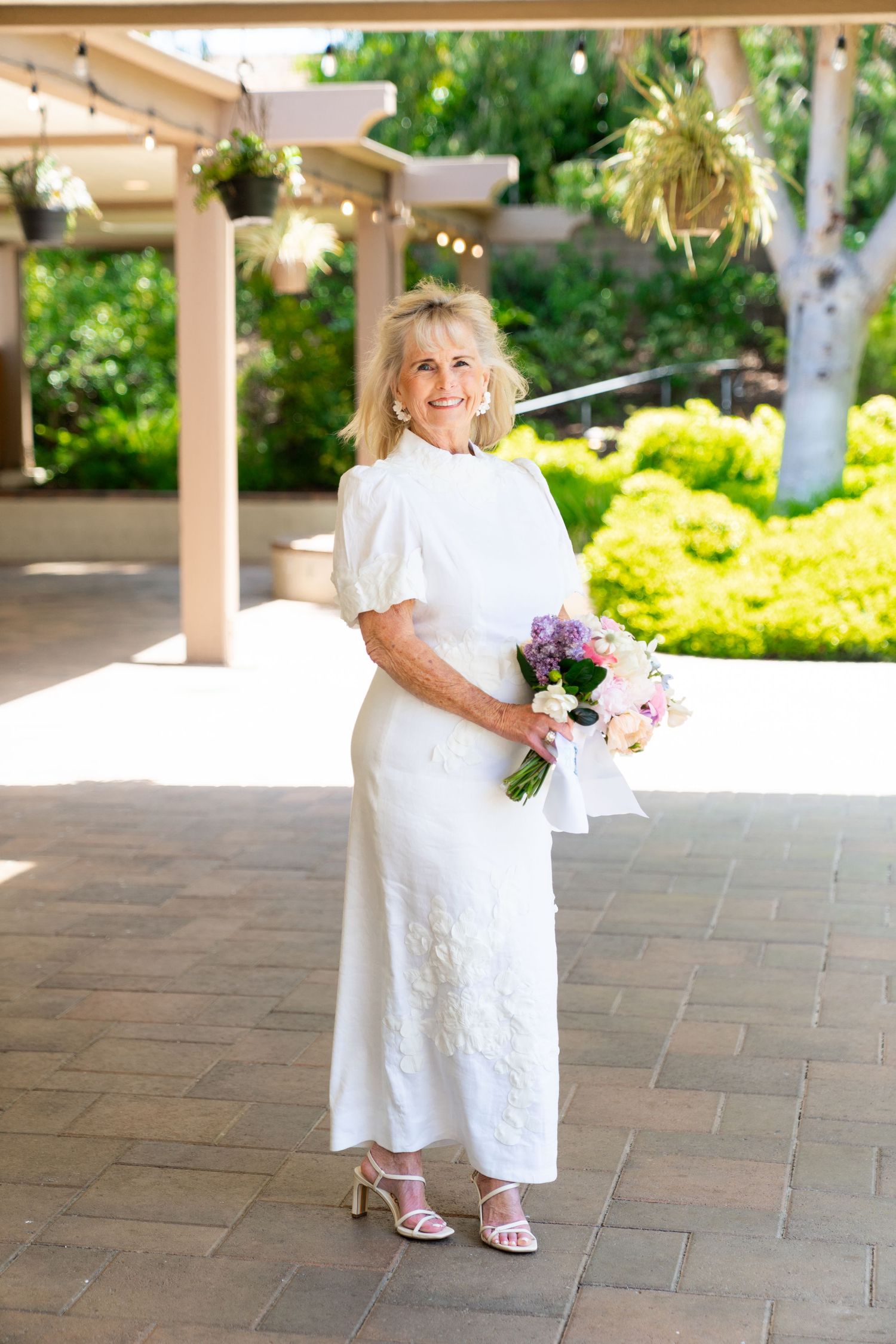 Elegant woman in white dress holding purple and pink bouquet on outdoor patio with garden backdrop.