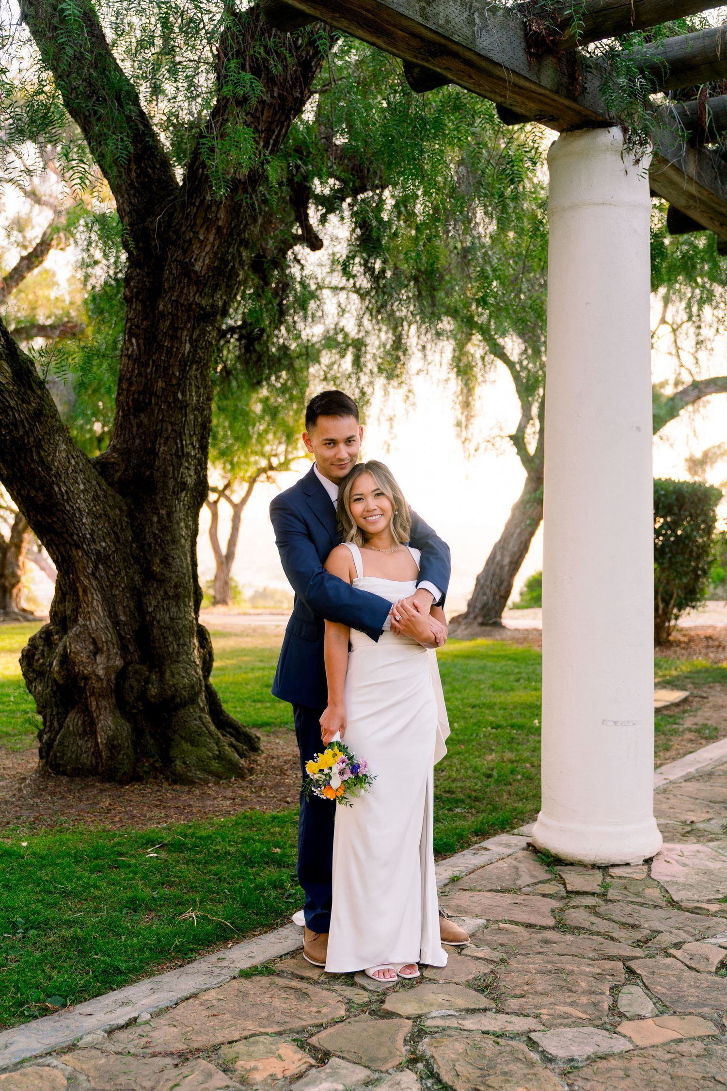 A couple in wedding attire embrace near a white column and old tree on stone path at sunset.