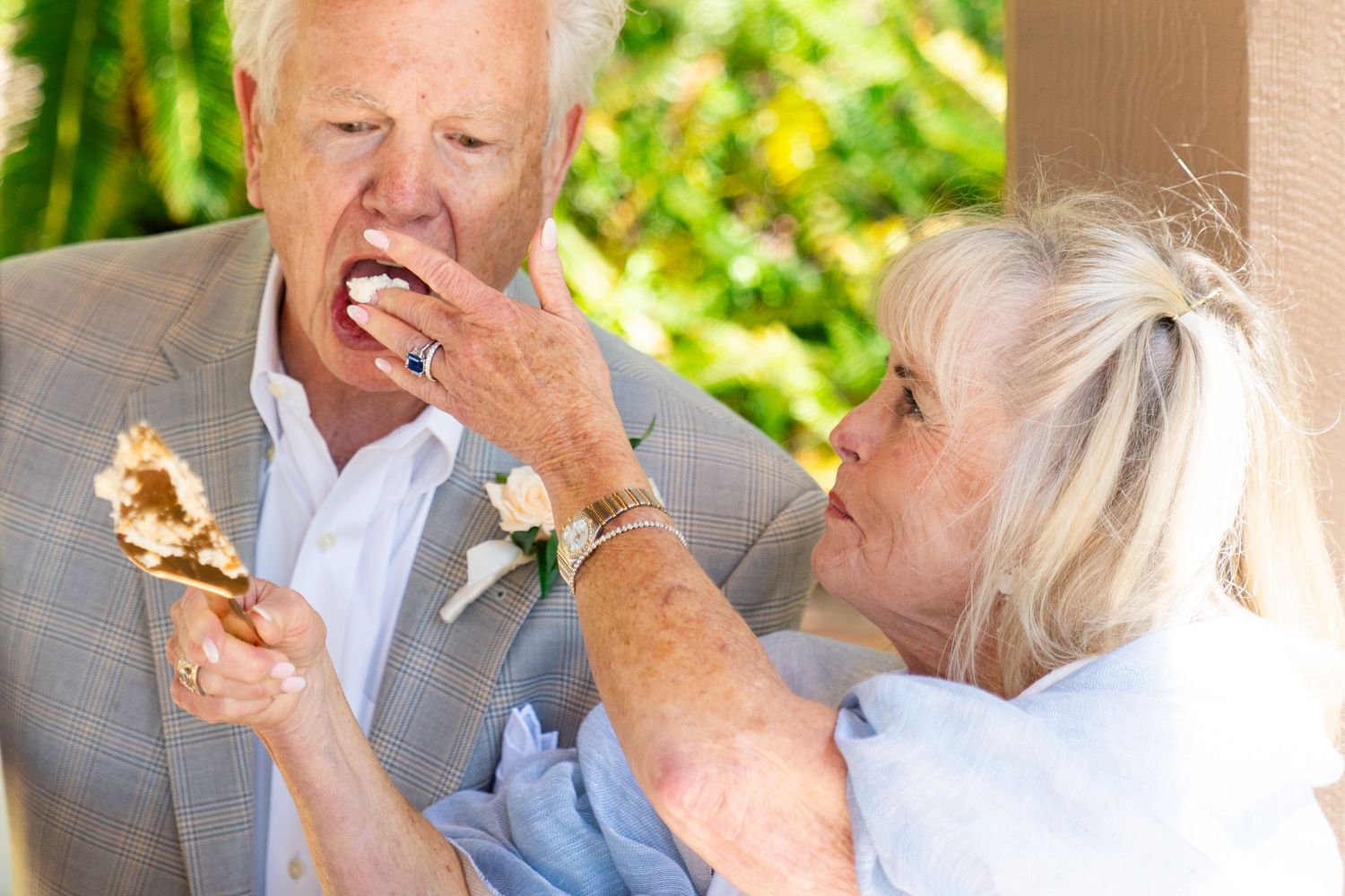 A senior couple in formal attire share wedding cake together outdoors on a sunny day.