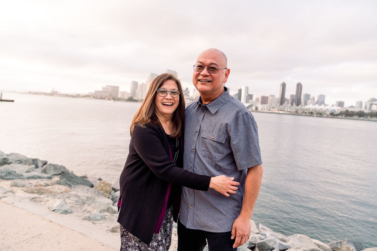 A couple stands by the waterfront with a city skyline view in the background.