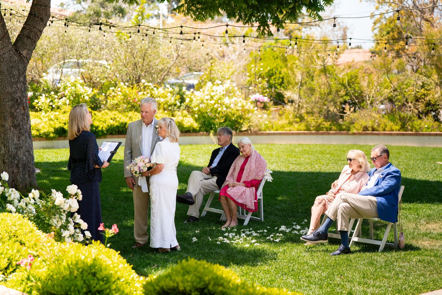 A small intimate outdoor garden wedding ceremony with guests seated on benches beneath a tree on a sunny day.