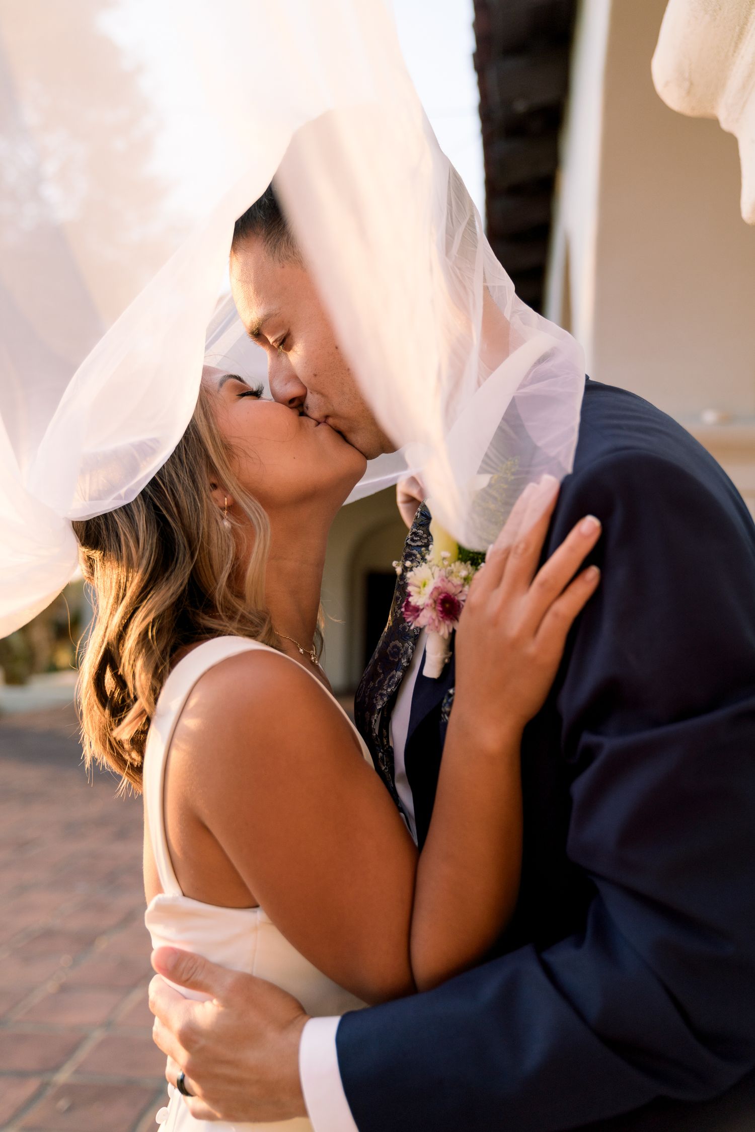 Romantic wedding kiss captured through flowing white veil in golden sunlight.