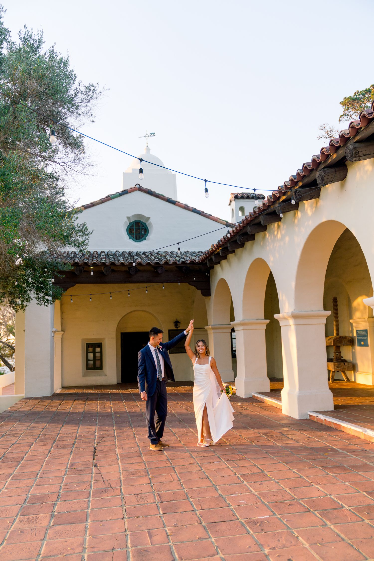 Wedding couple walking through Spanish-style courtyard with white arches and terracotta tiles.