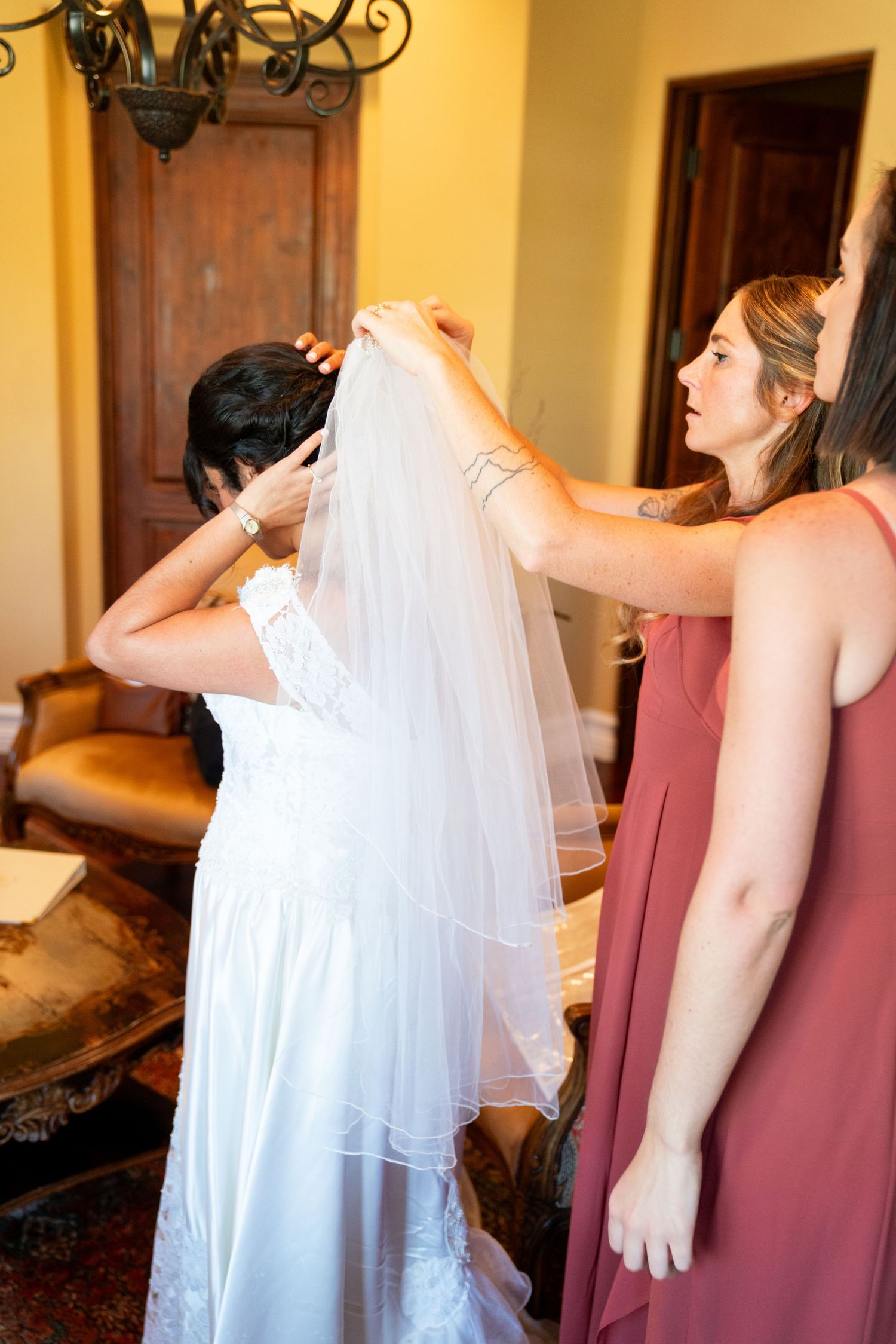 Bridesmaids help adjust a flowing white wedding veil during preparation in a warmly lit room.