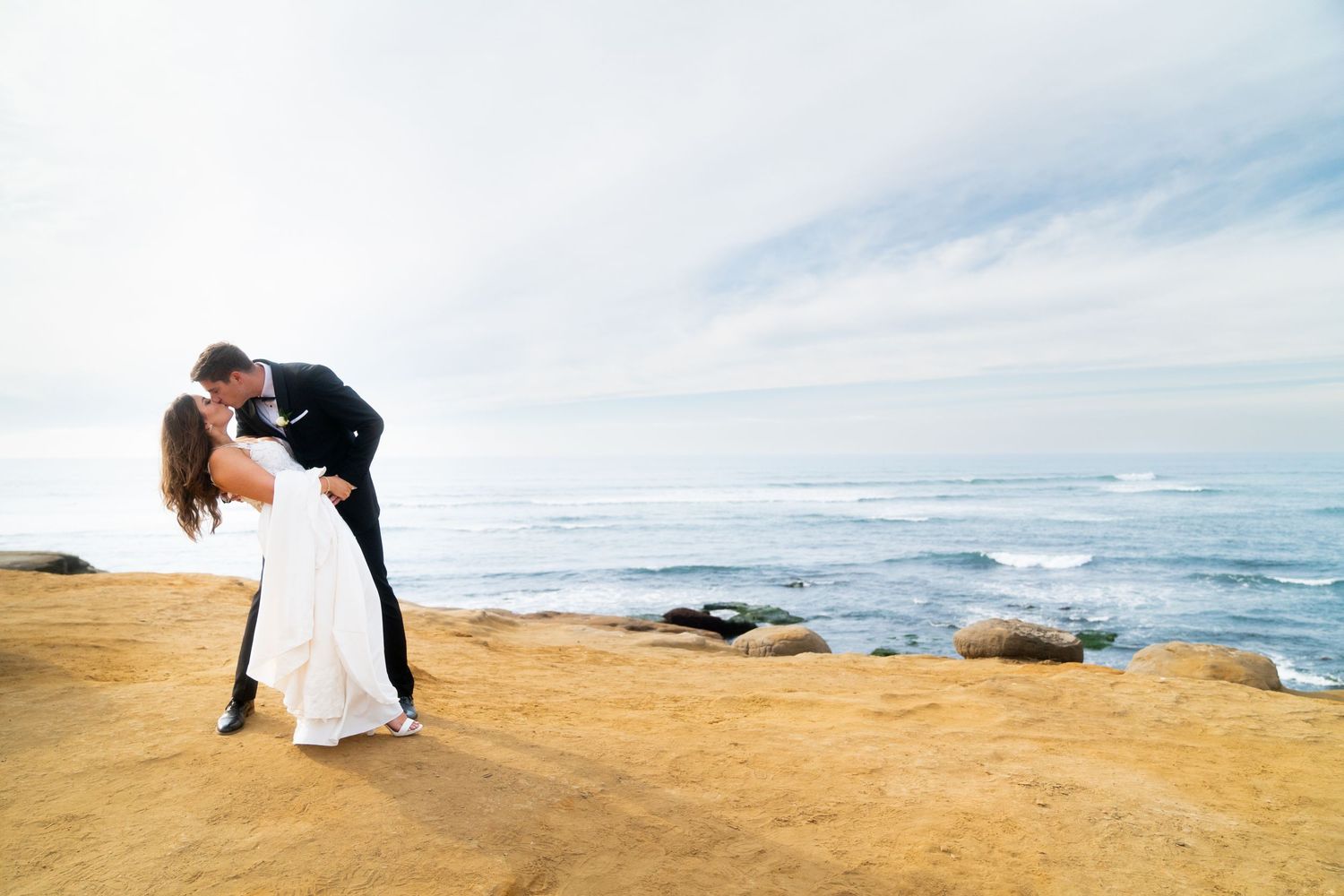 Two people in formal wedding attire embrace on a scenic rocky beach as waves crash against the shore at sunset.