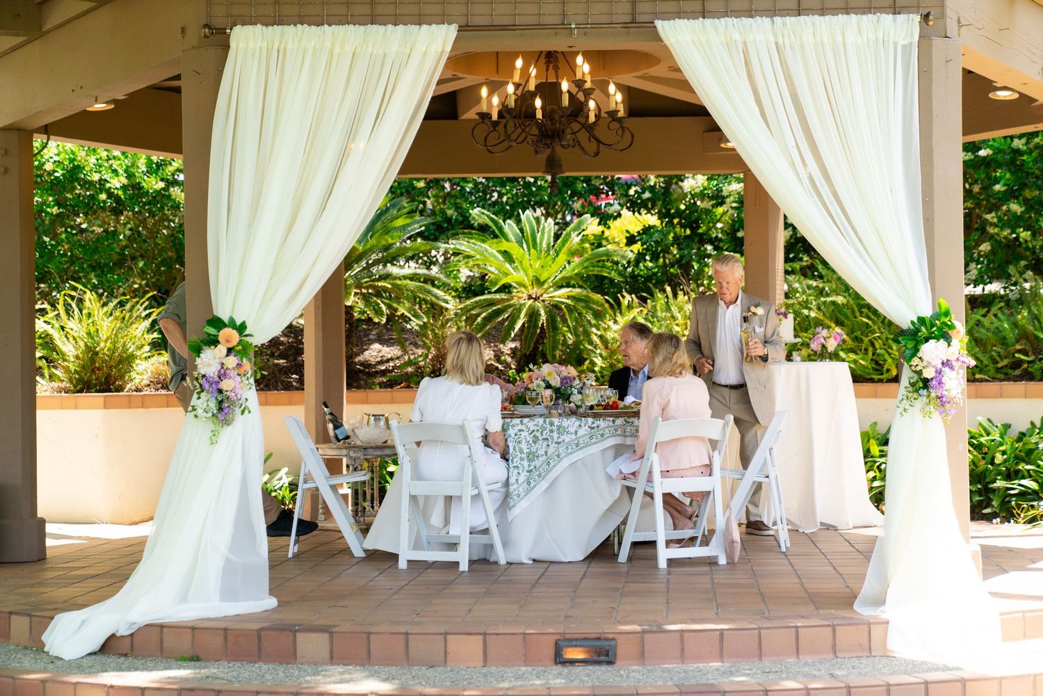 An elegant outdoor wedding ceremony setup under a covered pavilion with white drapes, chairs and floral arrangements.